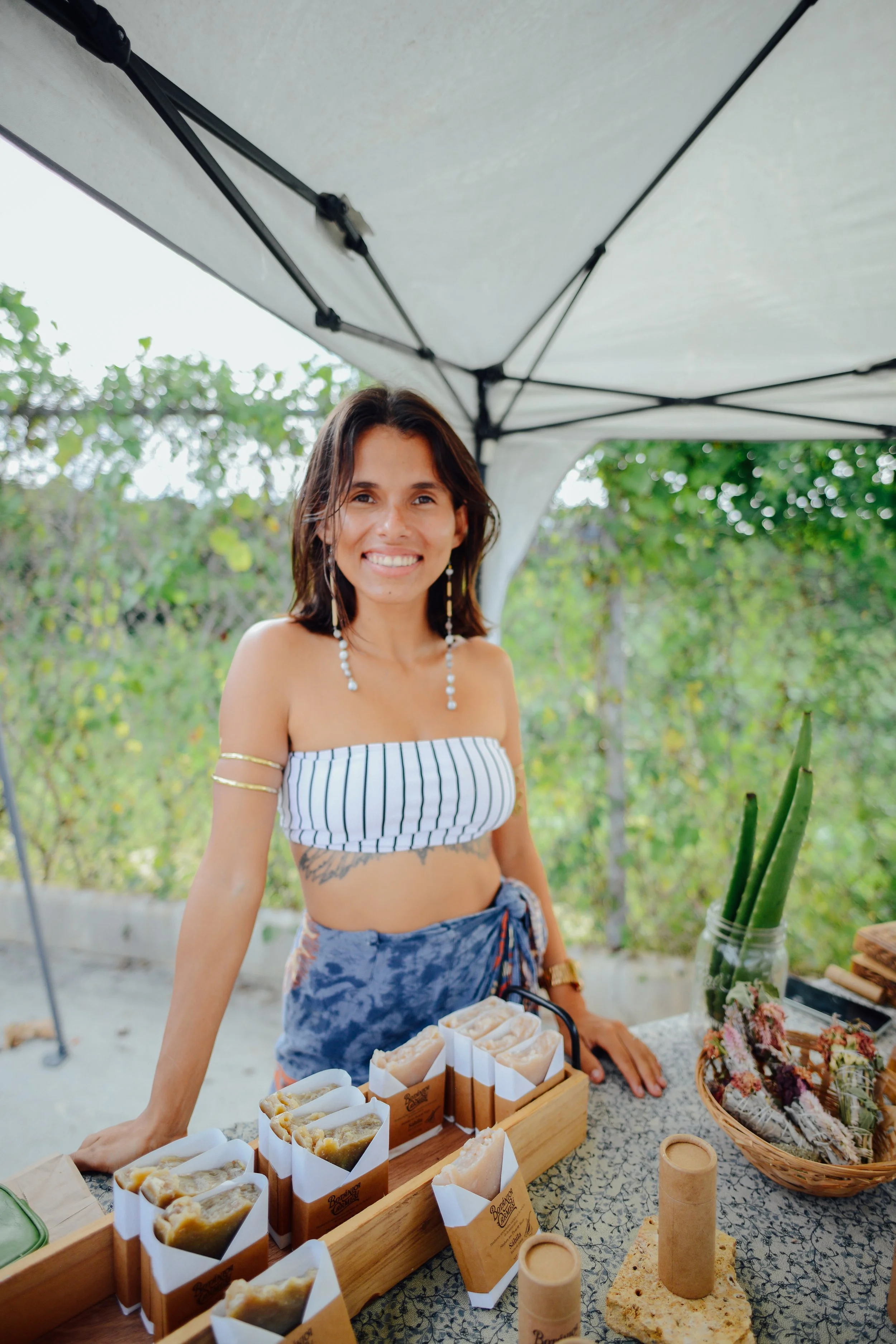 A smiling young woman standing behind a table displaying various handmade soaps at an outdoor market under a canopy, with greenery in the background.