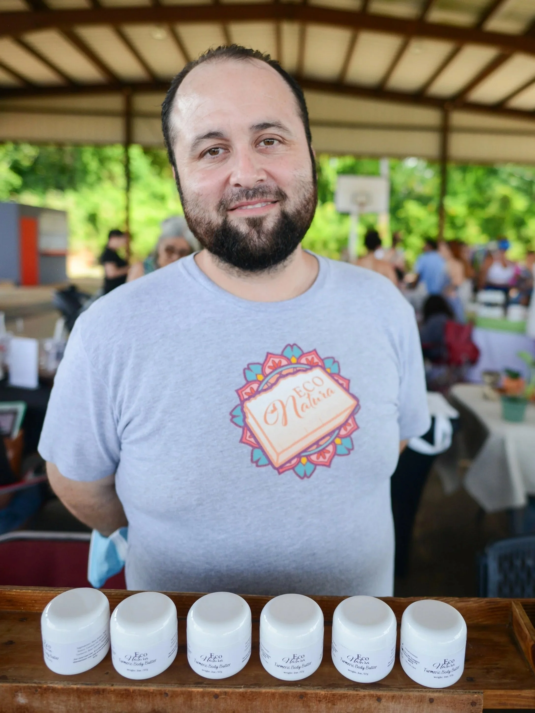 A man with a beard and mustache smiling at the camera, standing behind a row of jars labeled "Eco Native Turmeric Body Butter" at an outdoor event or market under a covered pavilion, with people in the background.
