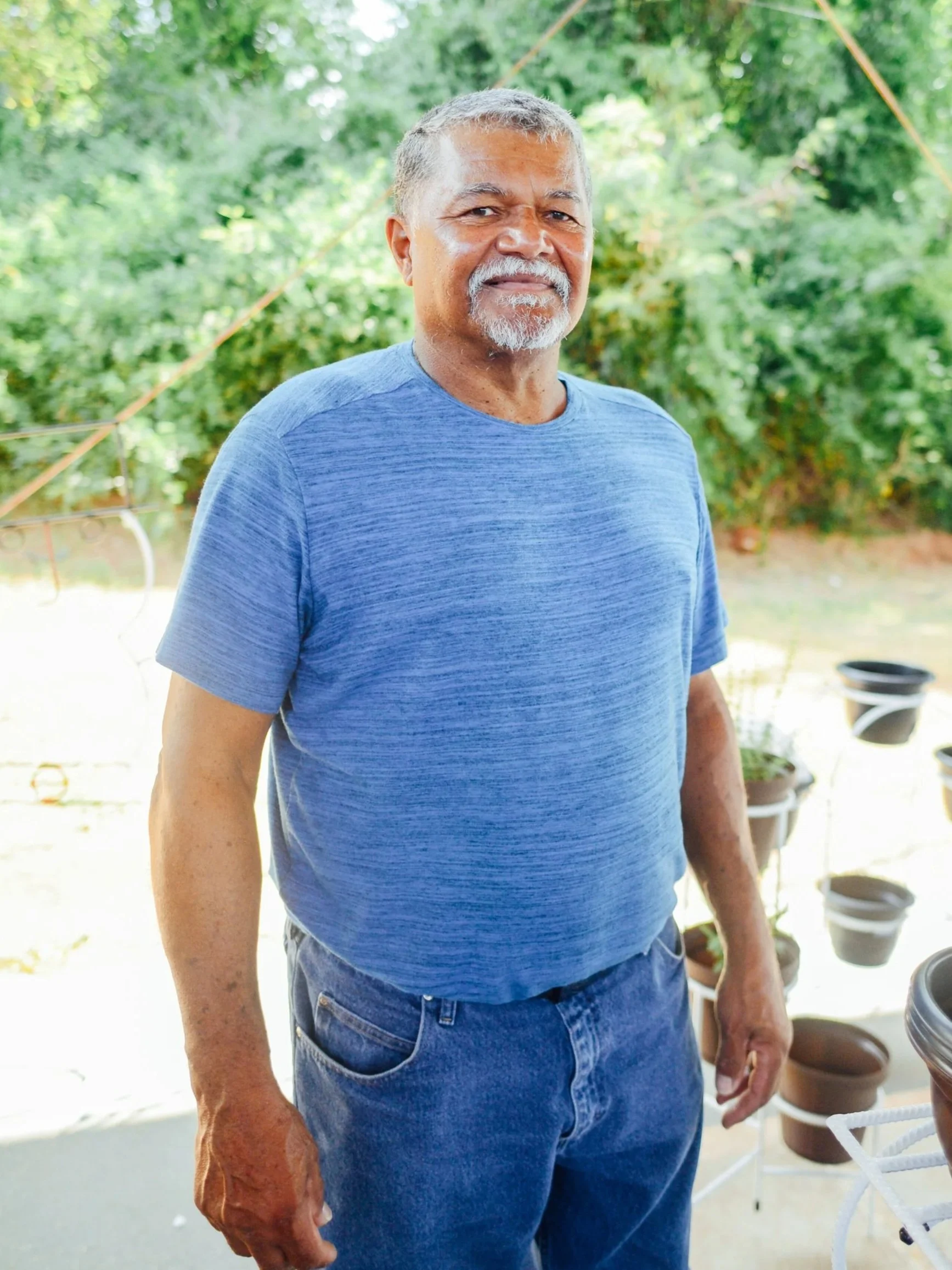 Older man with gray hair and beard wearing a blue t-shirt and jeans standing outdoors in front of green foliage.