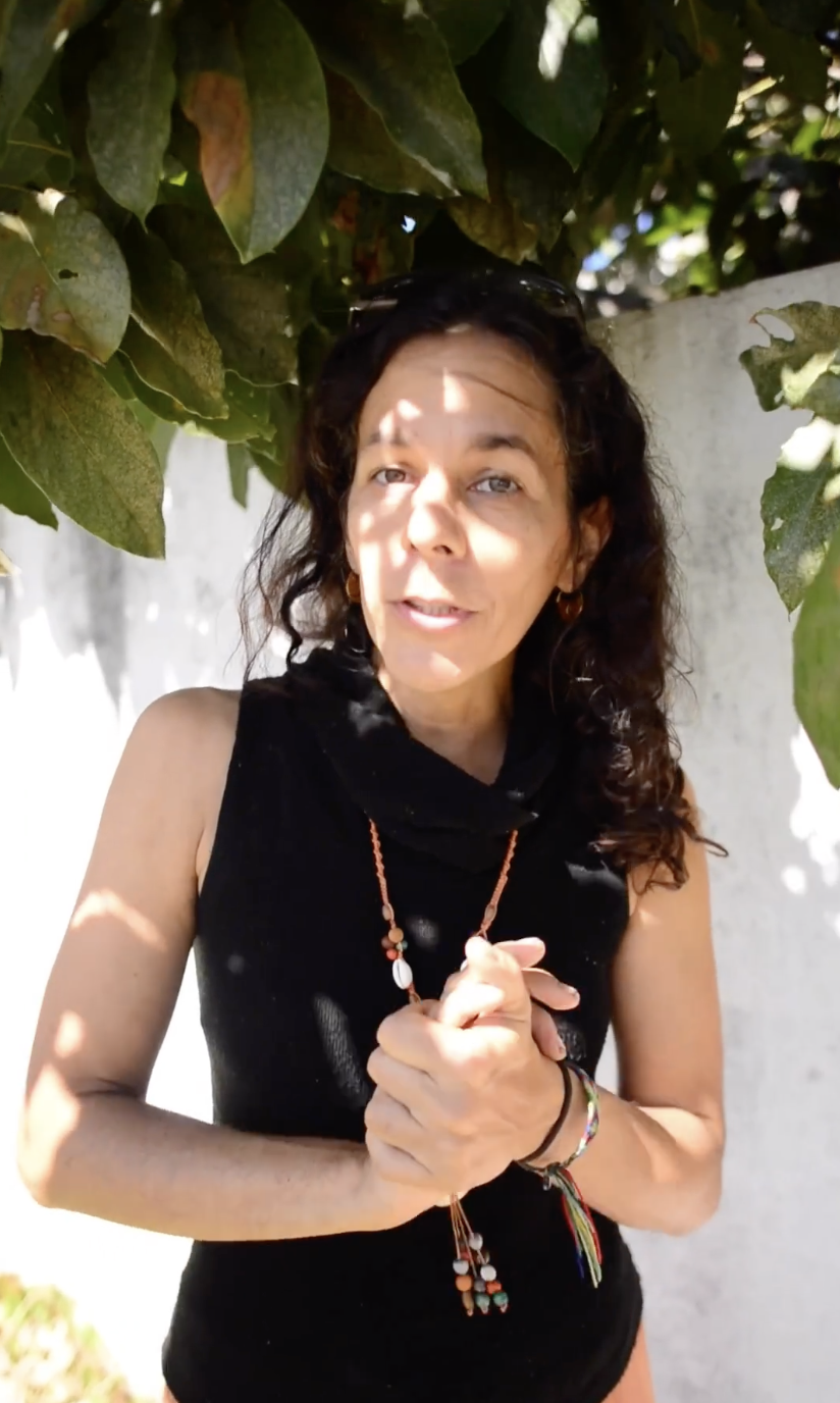 A woman wearing a black sleeveless top stands outdoors, shaded by tree leaves overhead, with light casting shadows on her face.