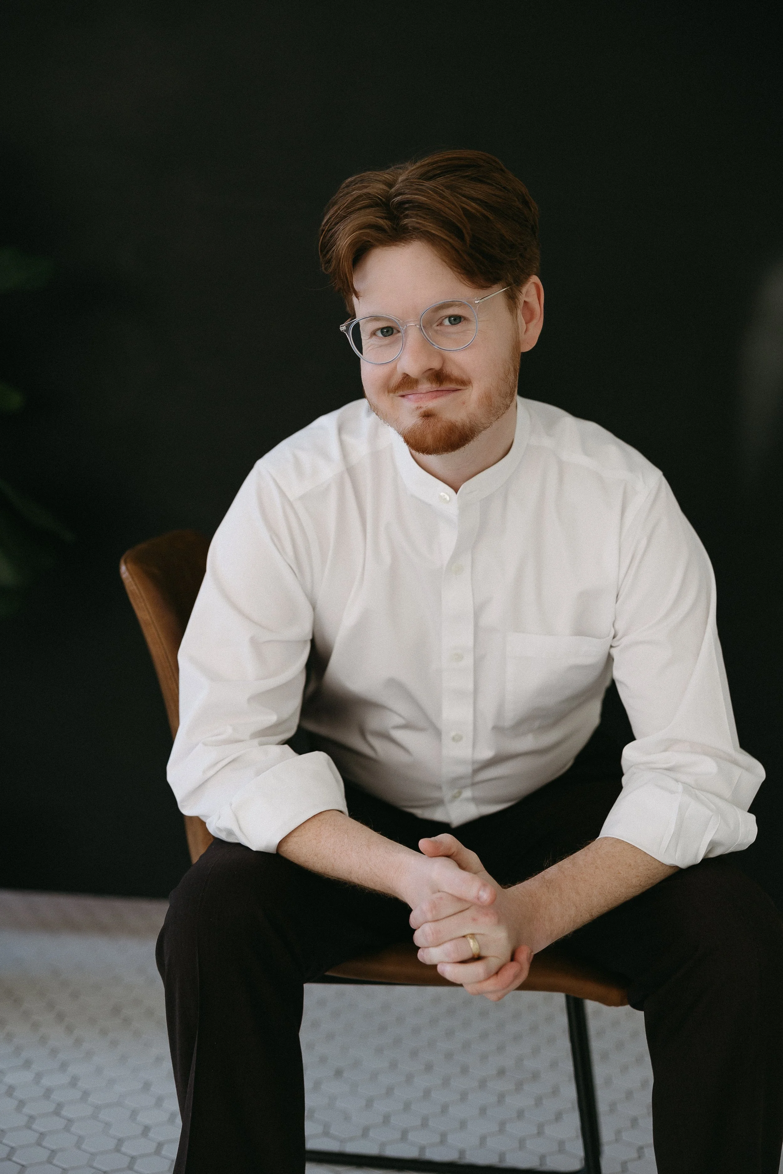 Dylon Crain, a man with reddish-brown hair, glasses, and a beard wearing a white button-up shirt, sitting on a wooden chair against a dark background.