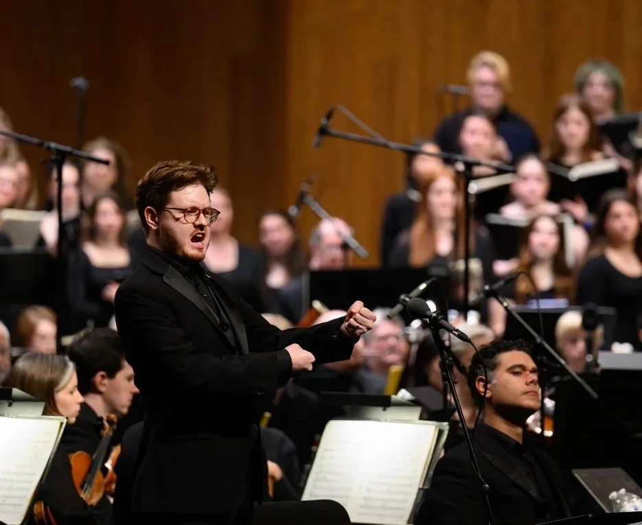 A man in a black suit and glasses is singing in front of an orchestra during a performance, with a choir in the background.
