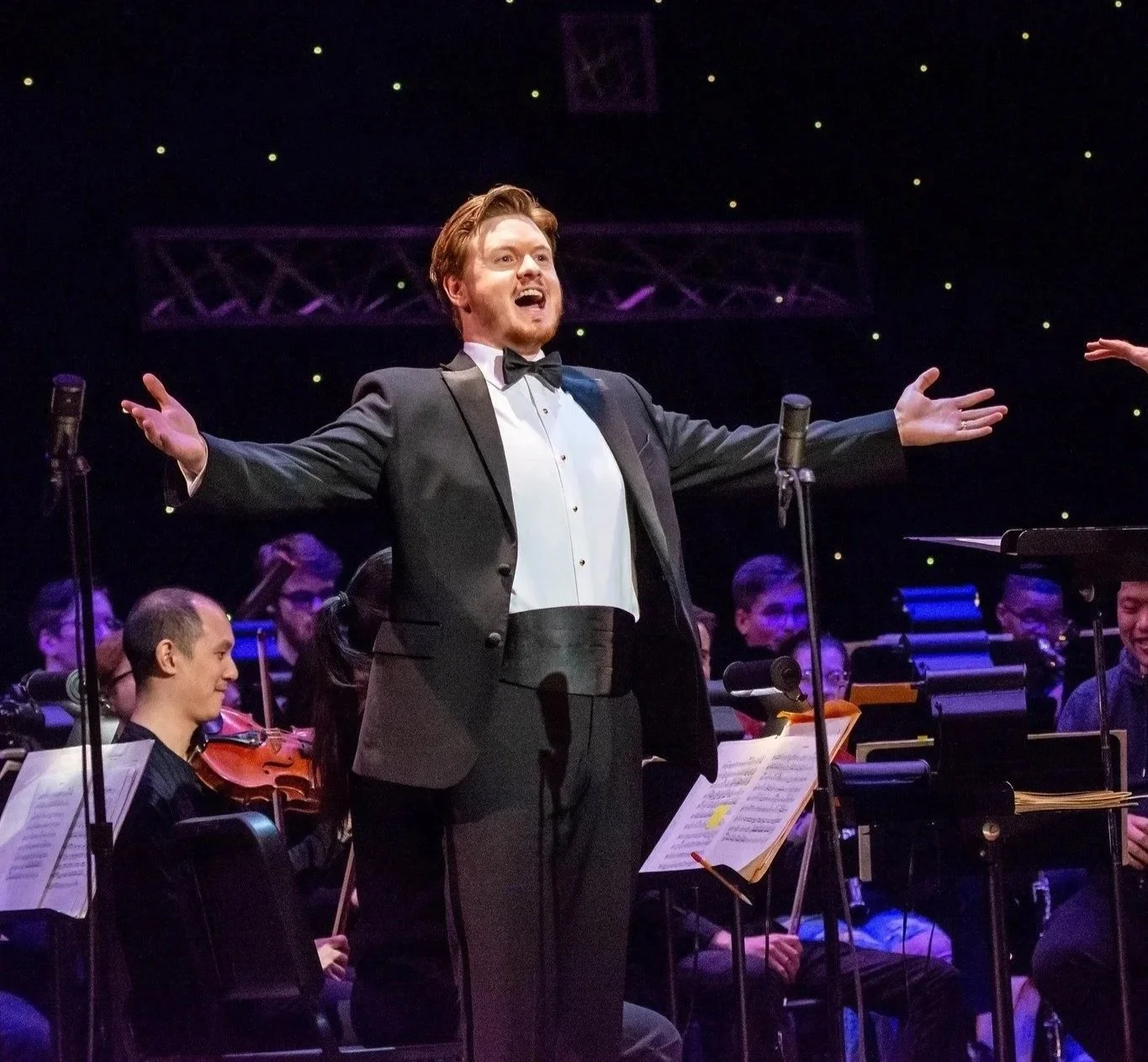 A male singer in a tuxedo performing on stage with an orchestra behind him.