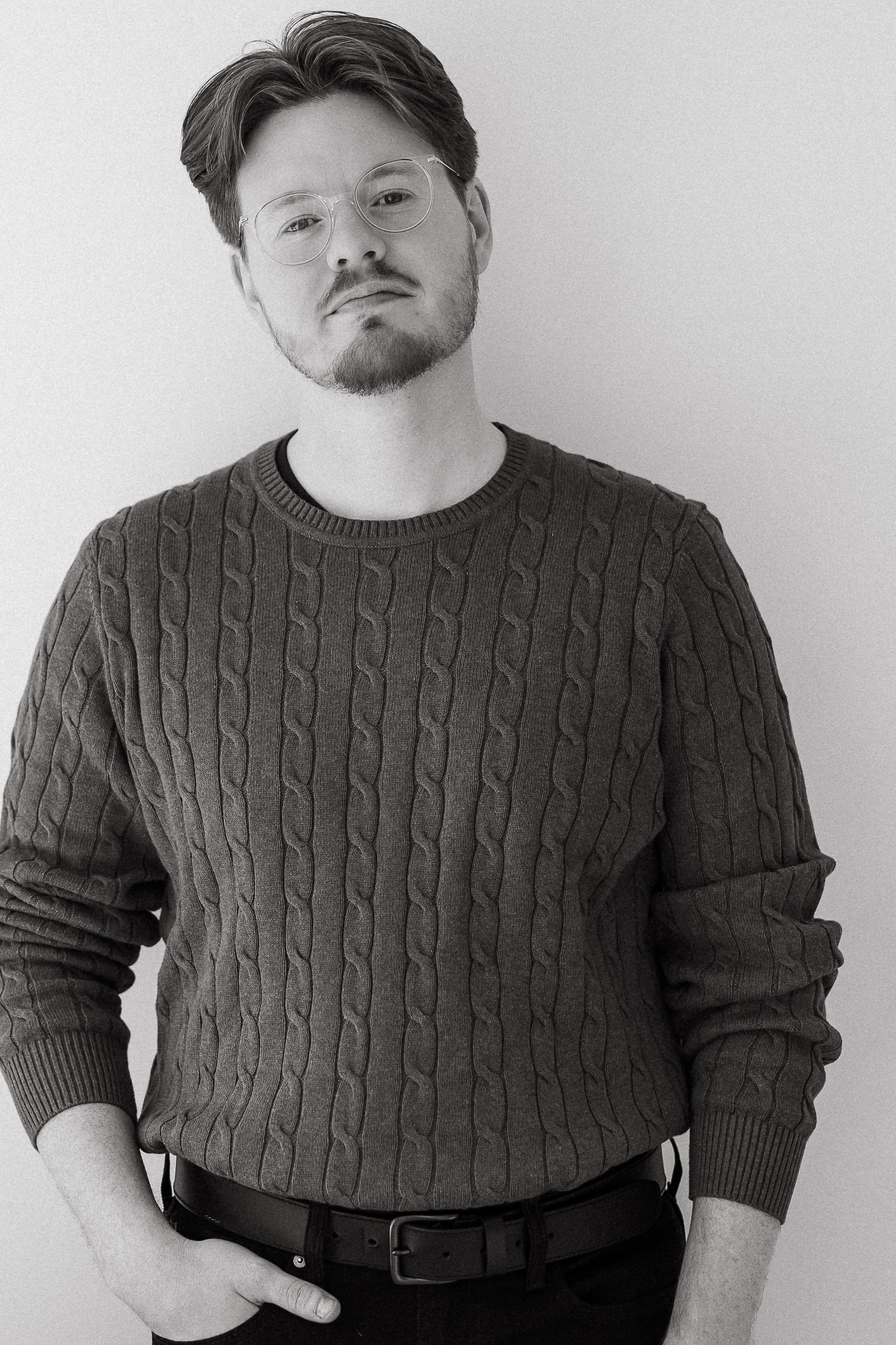 Black and white portrait of Dylon Crain, a young man with glasses, beard, and mustache, wearing a cable-knit sweater, standing with one hand in his pocket against a plain background.