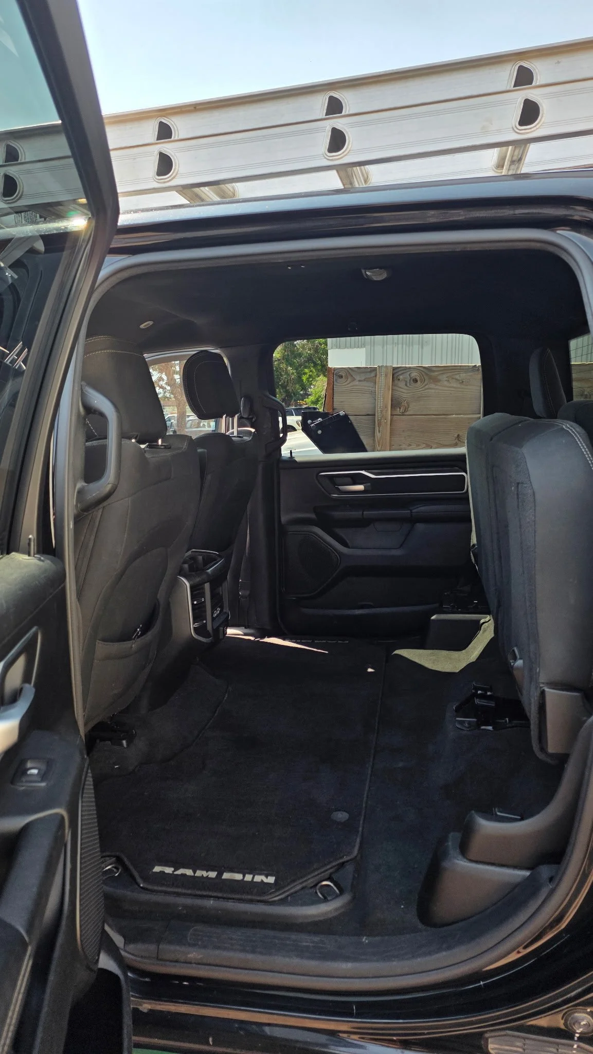 The image shows the interior of a black Ram pickup truck with the rear seats folded up, showing a view through the open rear passenger door. A wooden pallet is seen secured on the cargo area outside the vehicle, with a partial view of a building and 