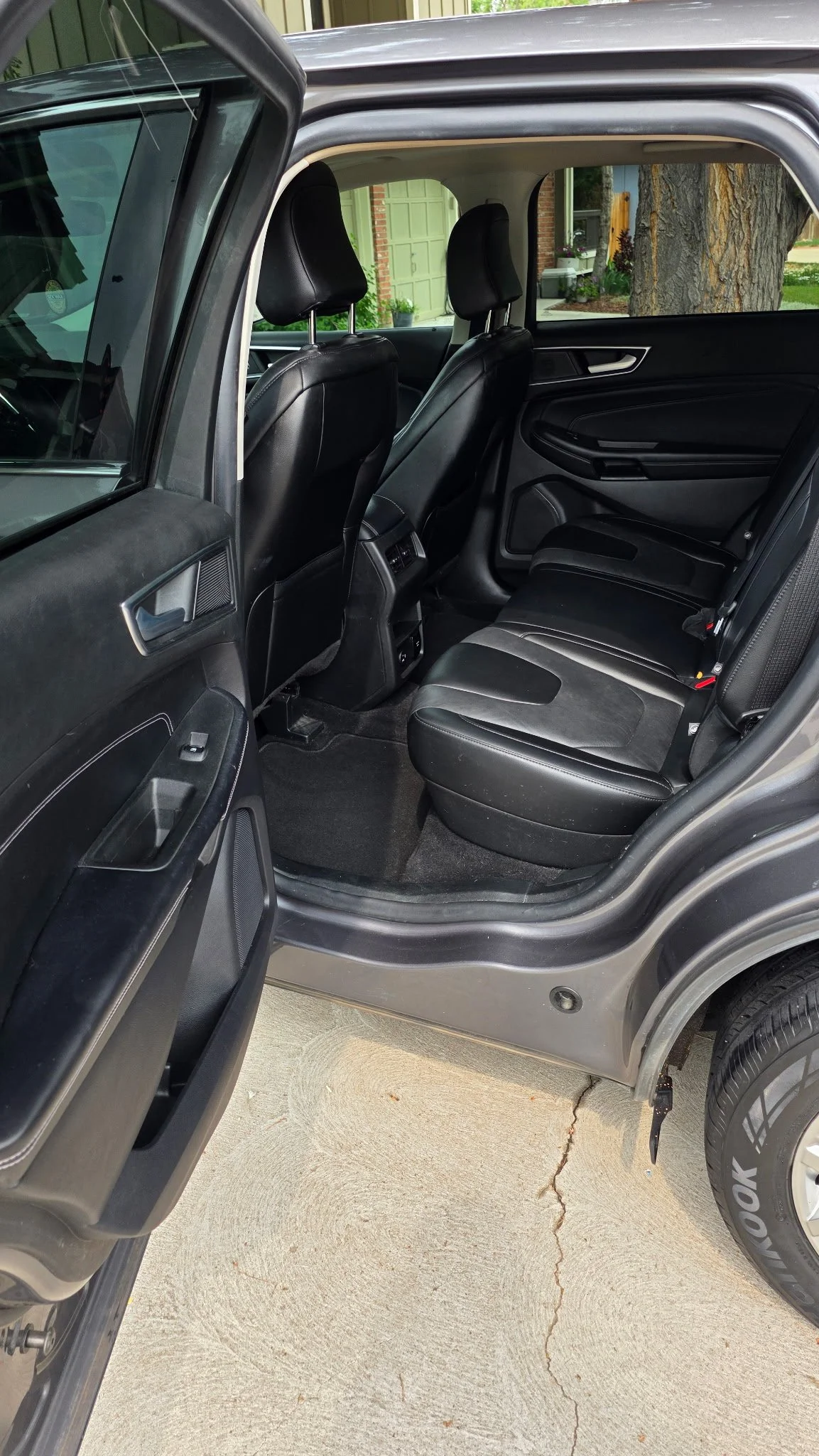 The interior of a silver SUV with black leather seats, shown with the rear passenger door open, parked on a concrete driveway with a large tree and house in the background.