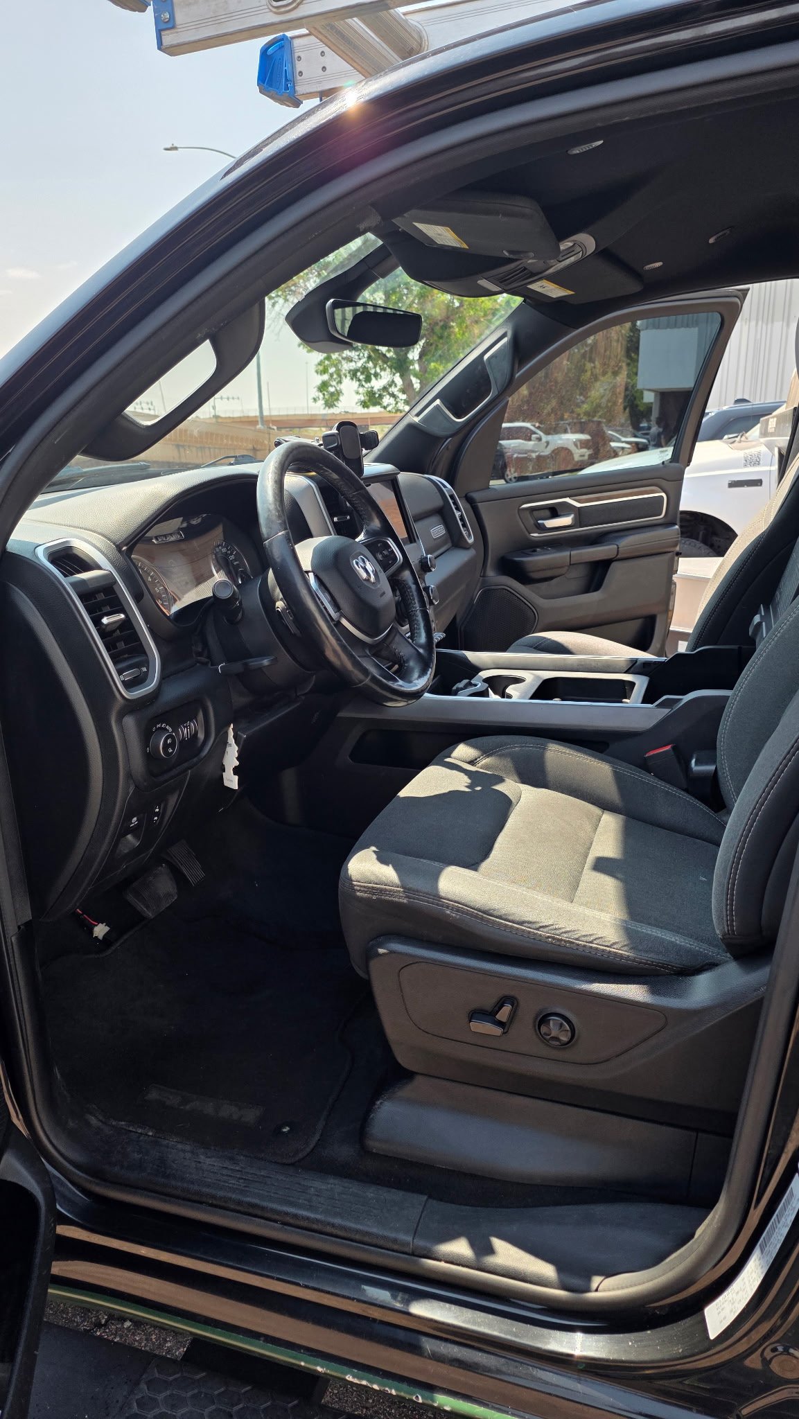 Interior of a pickup truck showing the driver and passenger seats, steering wheel, dashboard, and side door with window. Outside are other vehicles and trees.