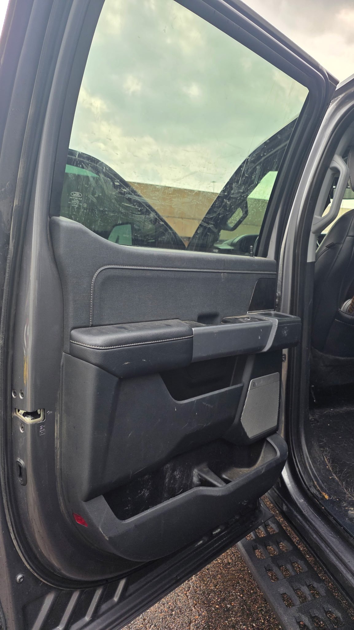 Inside view of a vehicle door showing window, door handle, and storage compartment, with another vehicle and cloudy sky visible through the window.