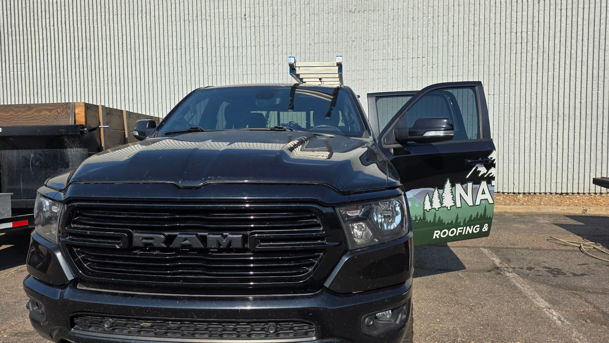 Front view of a black RAM pickup truck parked in front of a light-colored textured wall, with the passenger side door open revealing a partial view of the interior and a green and black sign on the door advertising roofing and insulation services.