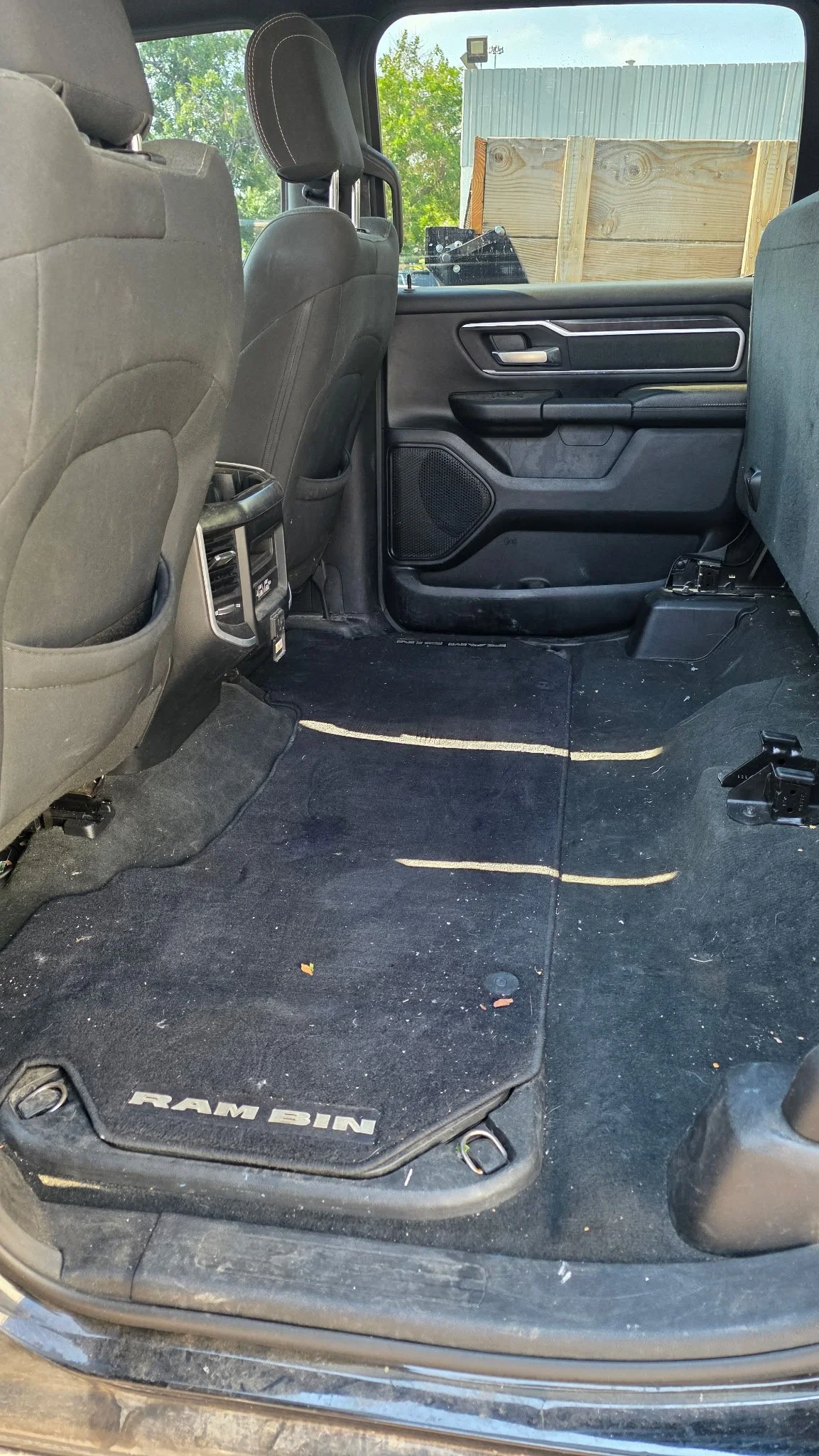 Rear interior of a Ram 1500 pickup truck showing black carpeted floor, Ram Bin floor mat, back of driver and passenger seats, and rear door on the passenger side with wooden crate outside the window.