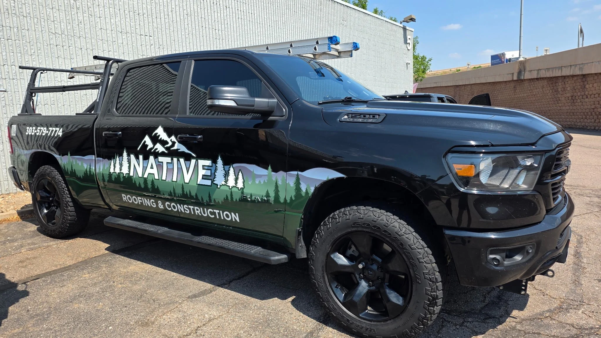 Black pickup truck with a mountain and forest graphic on the side, advertising a roofing and construction company, parked outside near a brick and metal wall, with a ladder mounted on the roof.