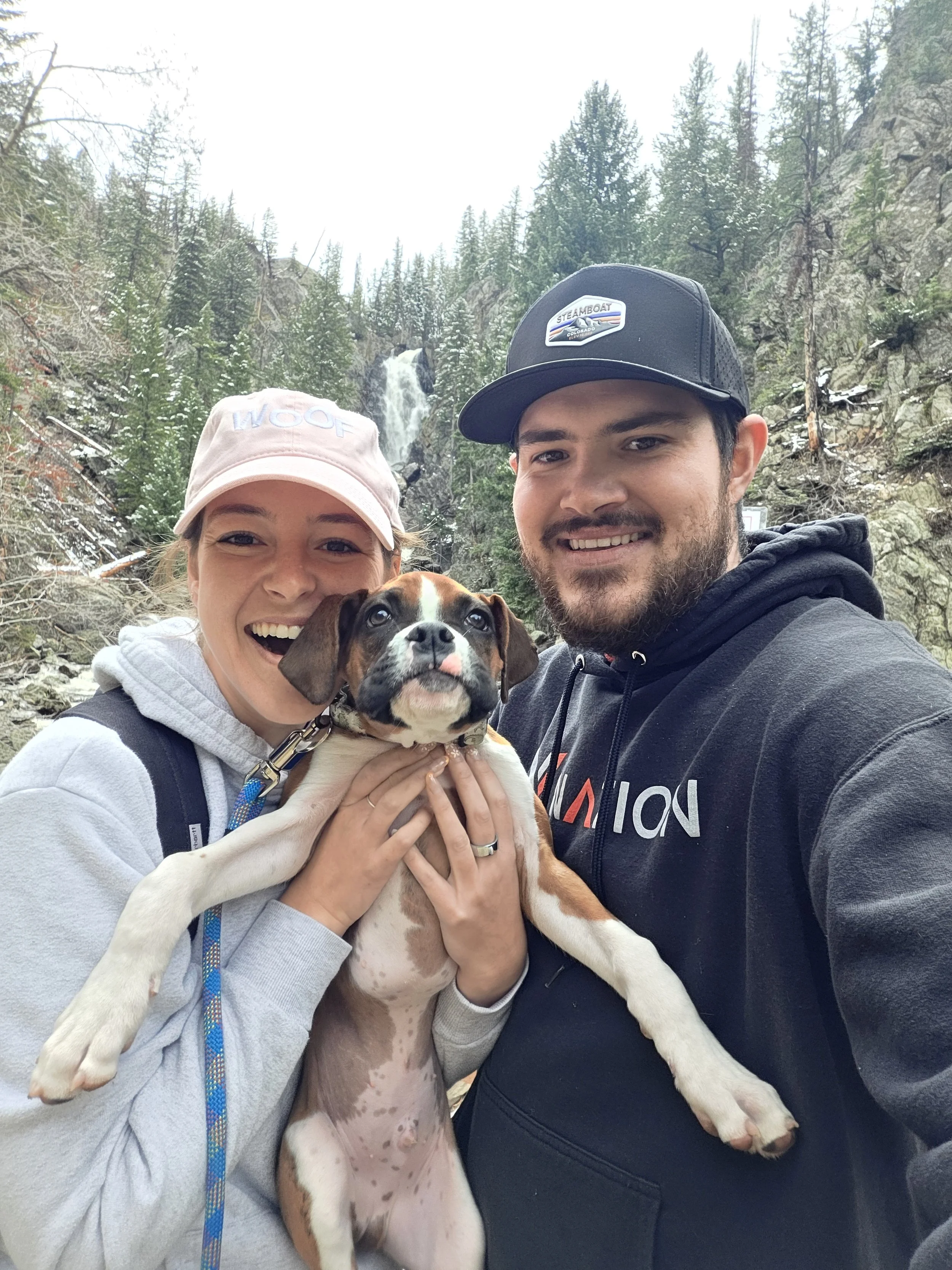 A happy couple with light skin holding a brown and white puppy outdoors in a rocky, forested area with trees and a waterfall in the background.