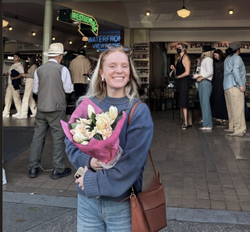 A young woman smiling and holding a bouquet of flowers outside a building with a group of people in the background.