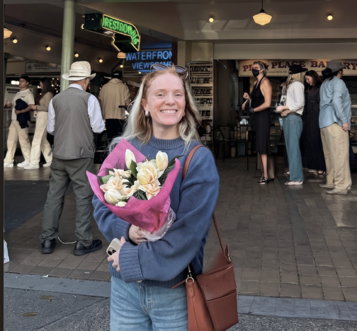 A young woman smiling and holding a bouquet of flowers in a public indoor space with other people in the background.