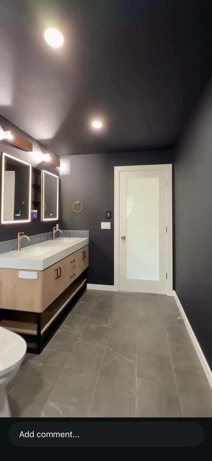 A modern bathroom with dark gray walls and ceiling, a double vanity with light wood cabinets and white countertops, gold fixtures, one mirror with LED lighting, gray floor tiles, and a white door with frosted glass.