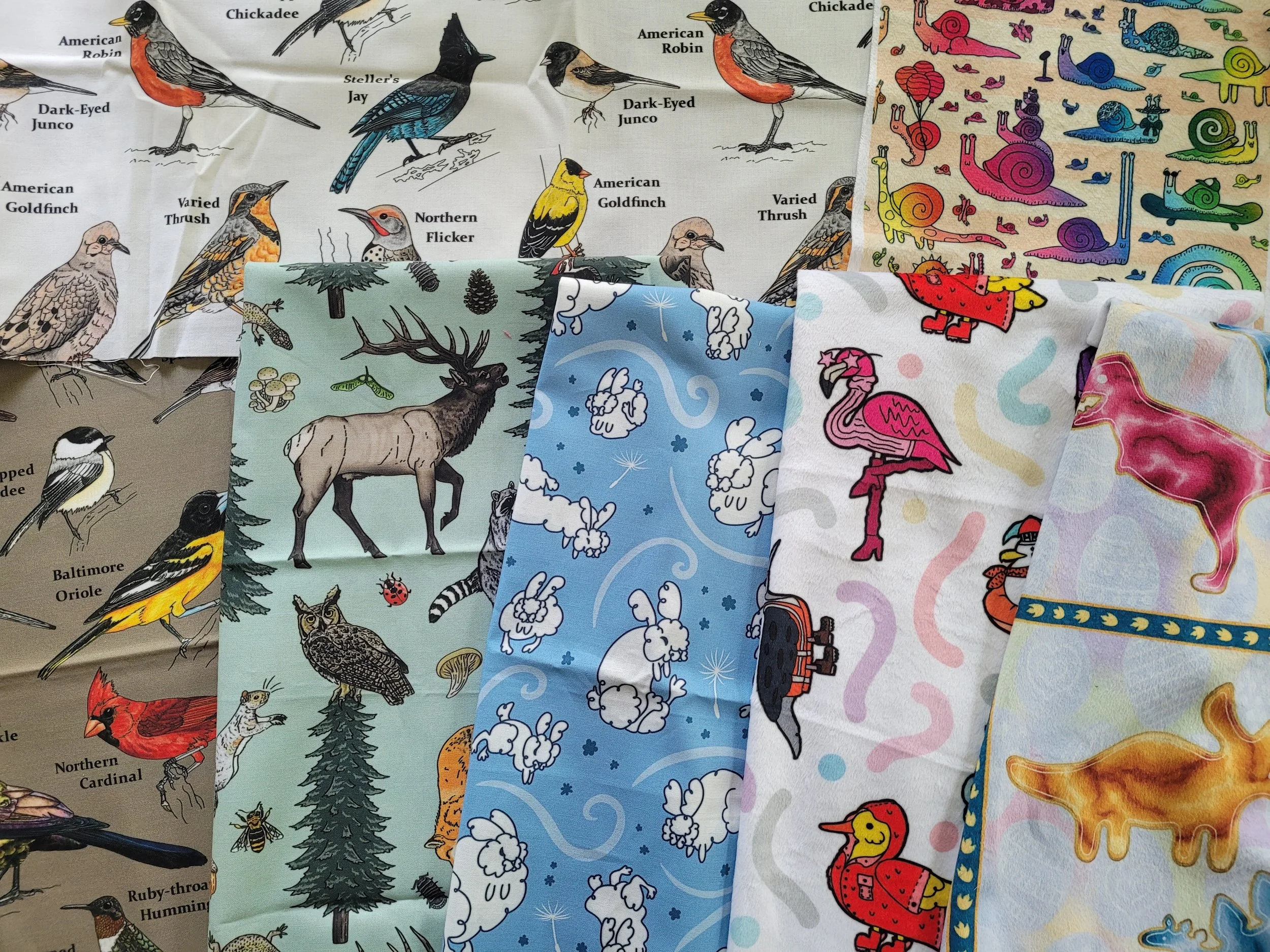 Various fabric swatches with my patterns on them, printed from Spoonflower. The fabrics all include animals or wildlife, from birds to elk, bunnies, and snails.