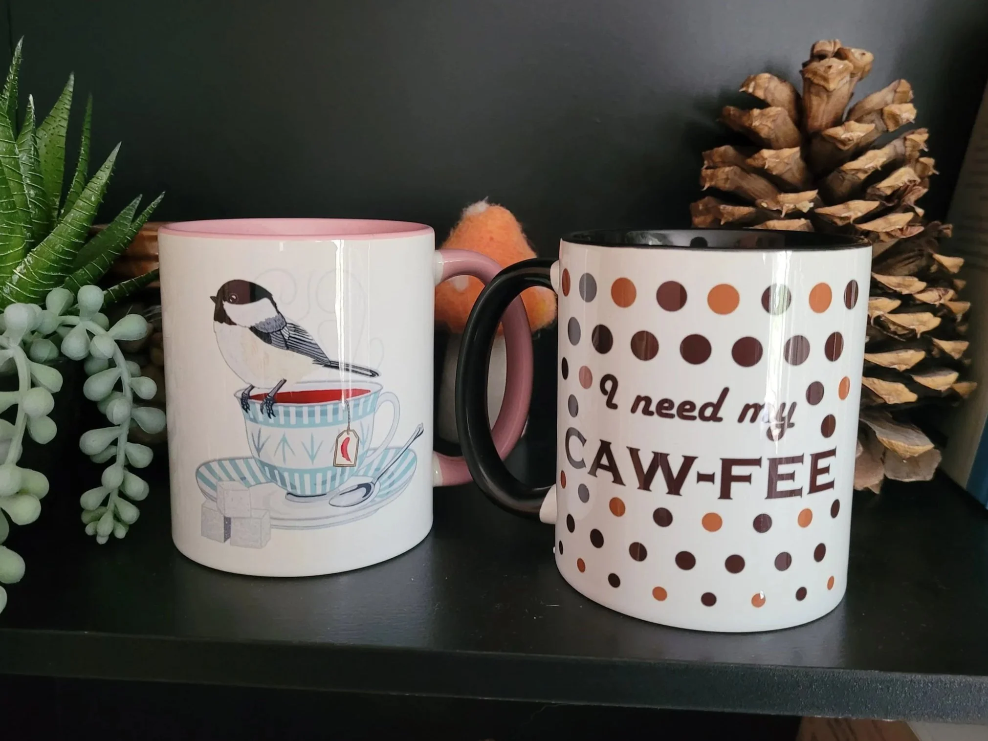Image of two mugs on a bookshelf. Left mug has pink accents and features a chickadee bird sitting on a teacup, right mug features the text "I need my CAW-FEE" surrounded by brown polka dots. Background contains plants and a pinecone.