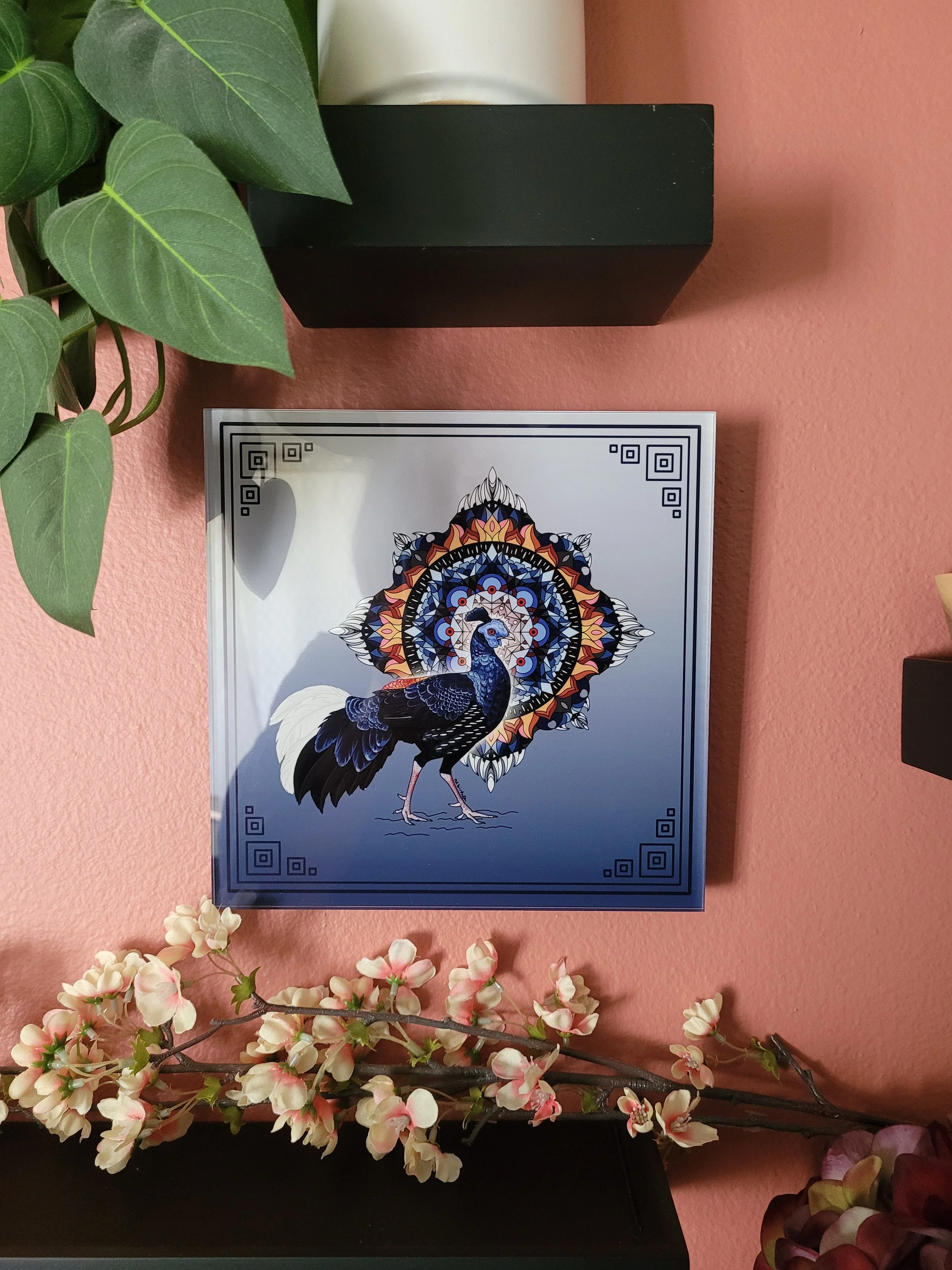 My artwork featuring a Malayan Crested Fireback bird with a radial, gemoetric design behind it. The piece is printed on an acrylic block, from INPRNT, and hanging on an orange wall. It's surrounded by black shelves and plants.