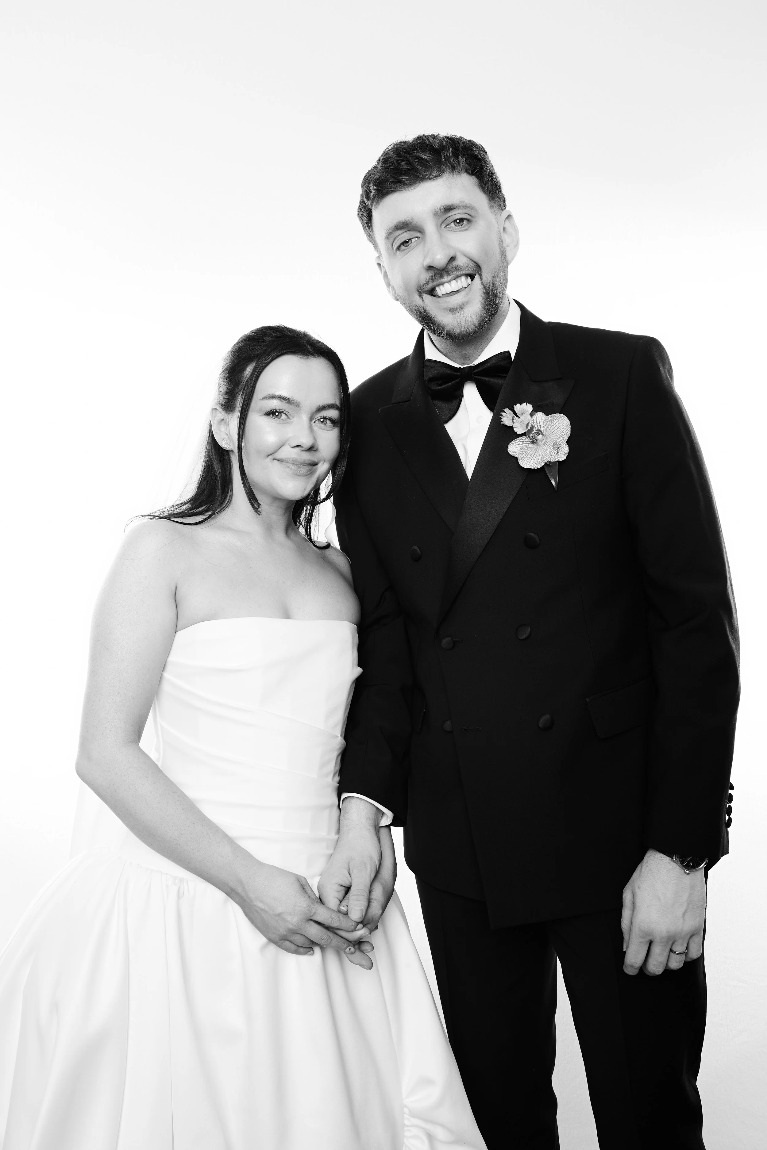 A bride and groom in wedding attire, smiling and holding hands, in a black-and-white portrait. The bride in a strapless white gown, the groom in a tuxedo with a bow tie and boutonniere.