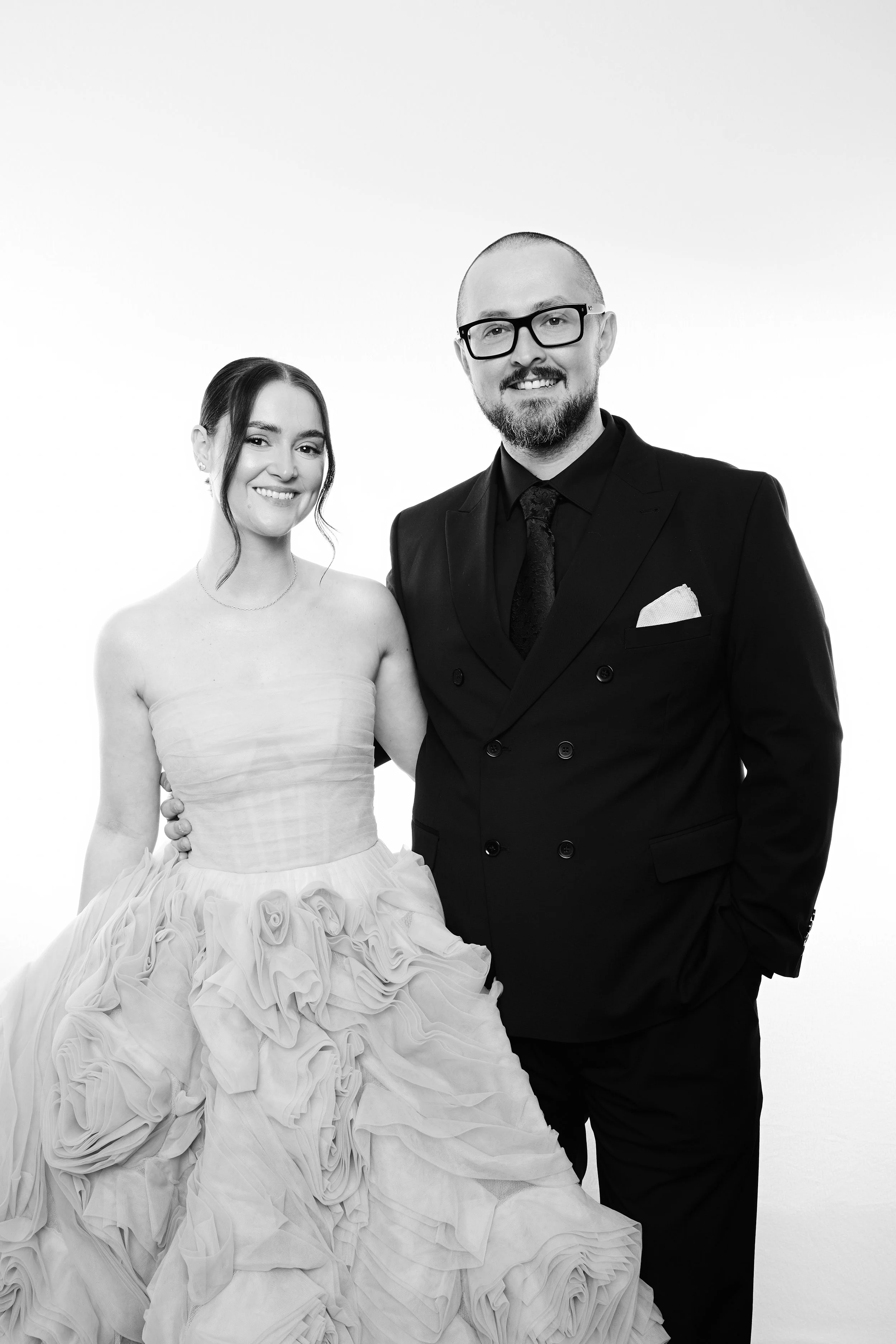 A black and white photo of a smiling couple, a woman in a strapless gown with ruffled layers and a man in a black suit with glasses, posing together against a plain background.
