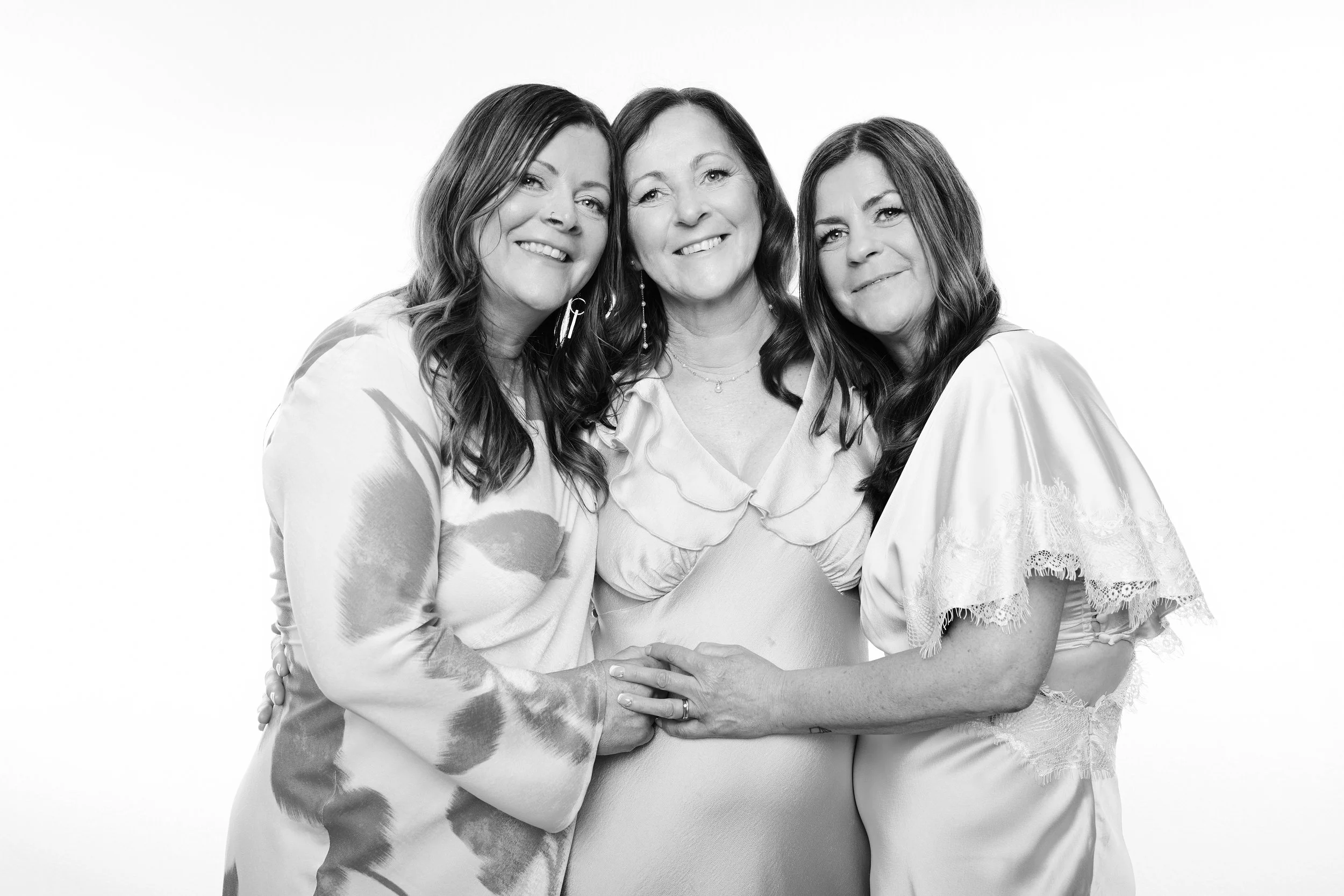 Black-and-white photo of three women embracing and smiling, standing in front of a plain background.