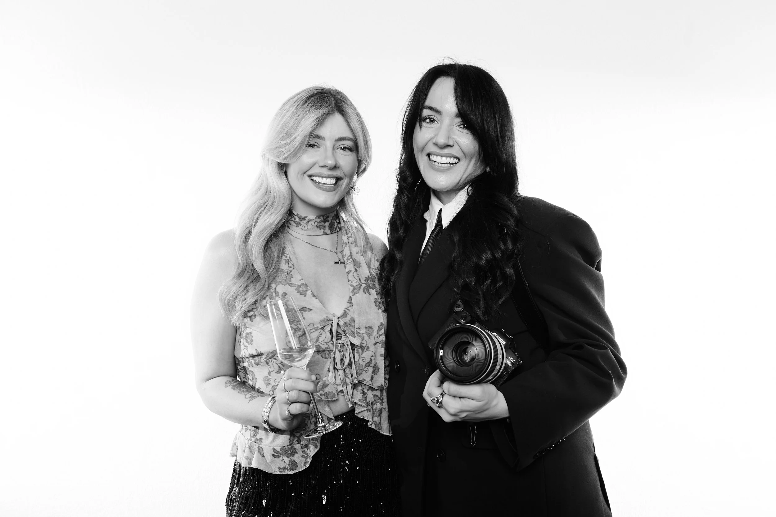 Two women smiling at a camera, holding a glass of champagne, in front of a plain white background.