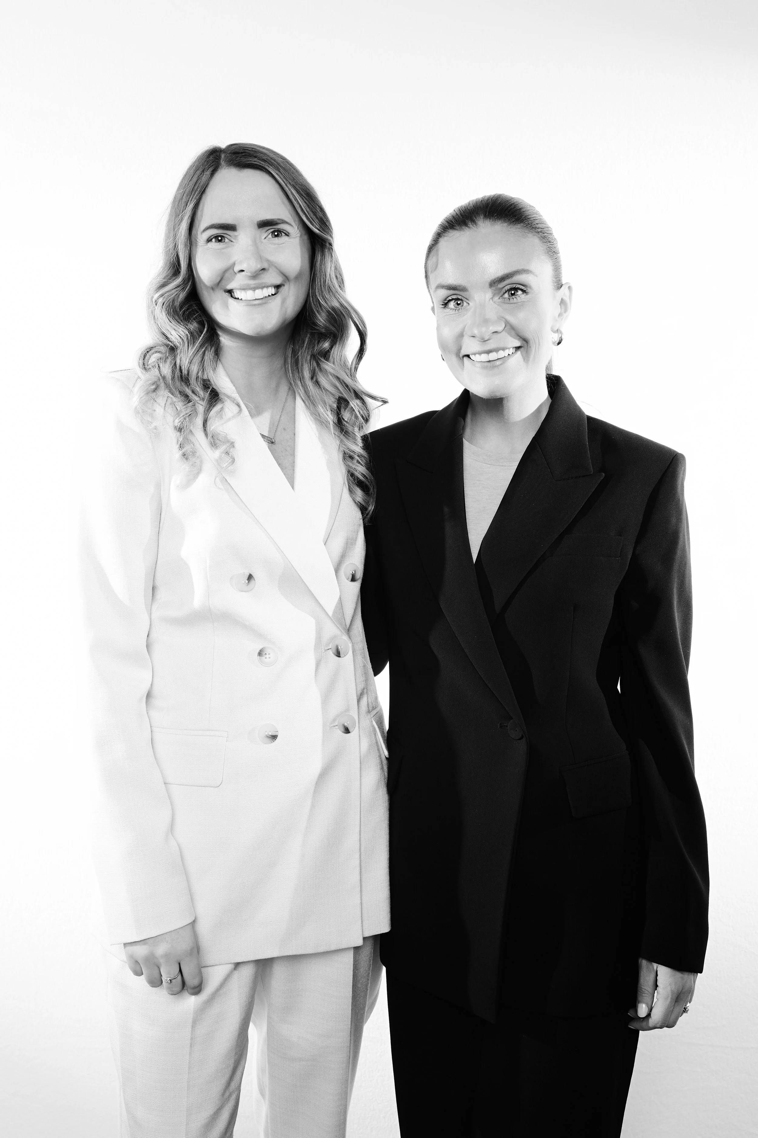 Two women standing side by side, smiling, in professional attire, with a plain white background.