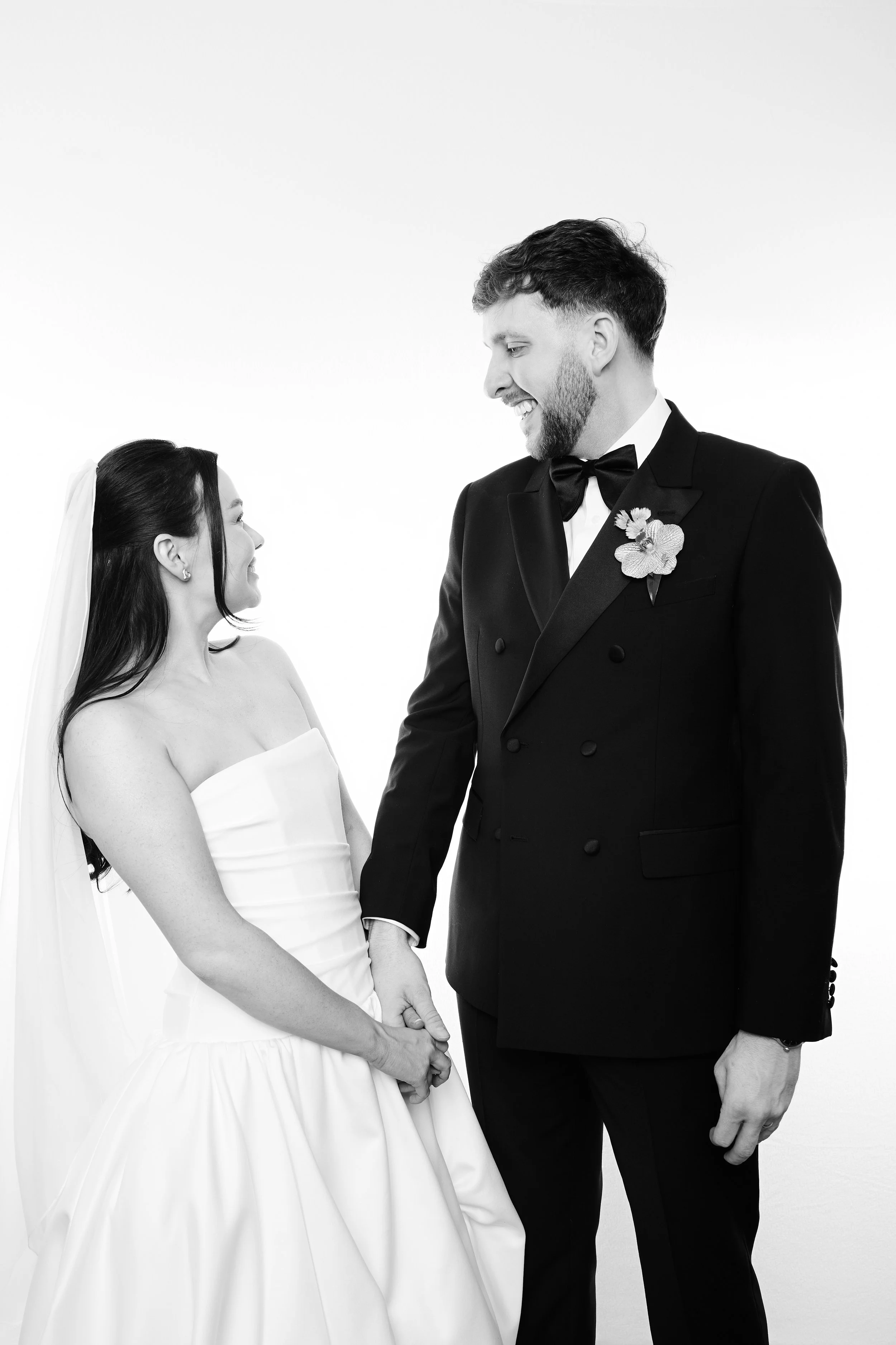 A black-and-white photo of a bride and groom holding hands, smiling at each other, standing close together against a plain white background. The bride is wearing a strapless wedding gown with a veil, and the groom is in a tuxedo with a boutonniere.