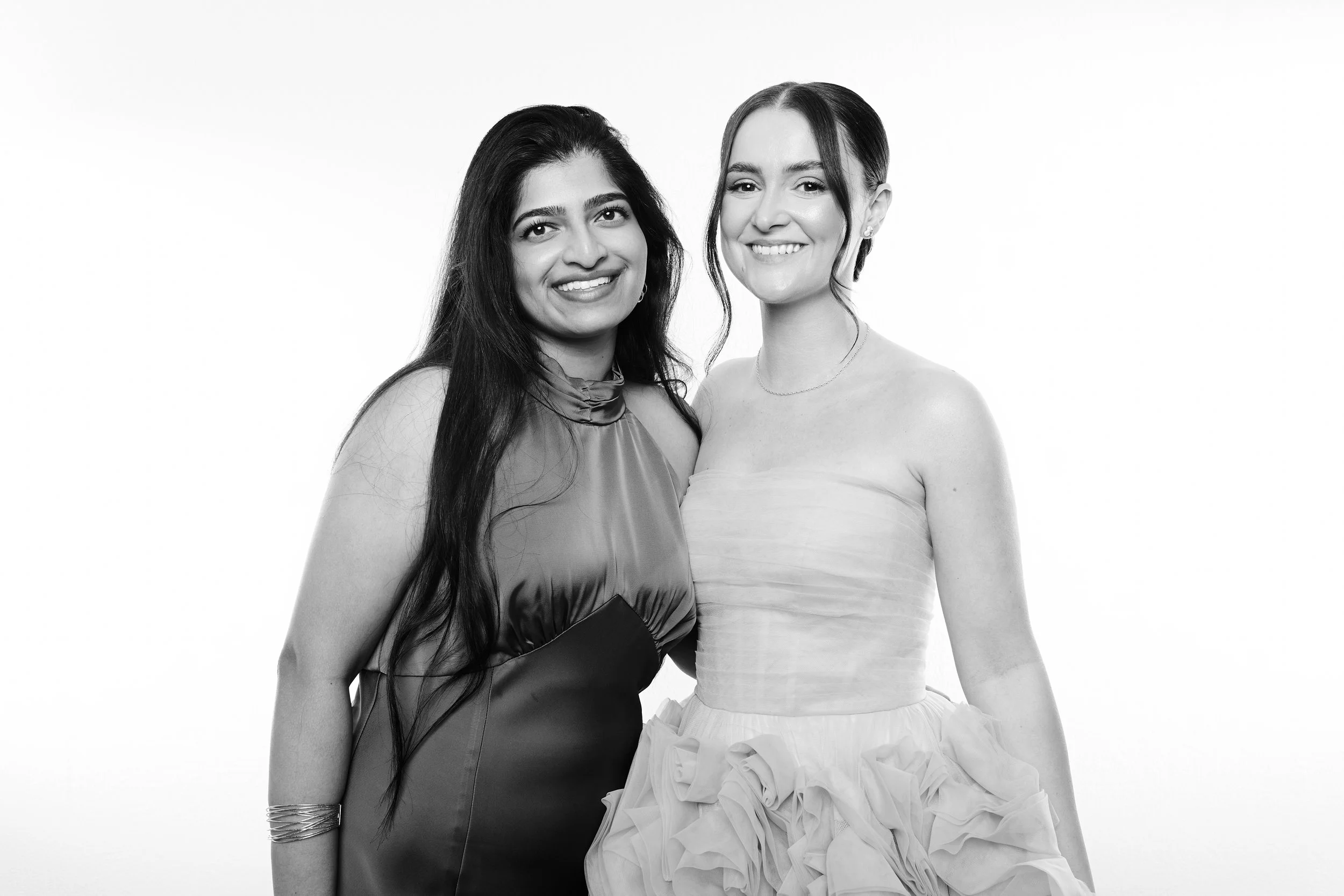 Two women standing close together, smiling, in black-and-white. One has long dark hair and is wearing a satin dress, the other has dark hair in an updo and is dressed in a strapless dress with ruffled skirt.