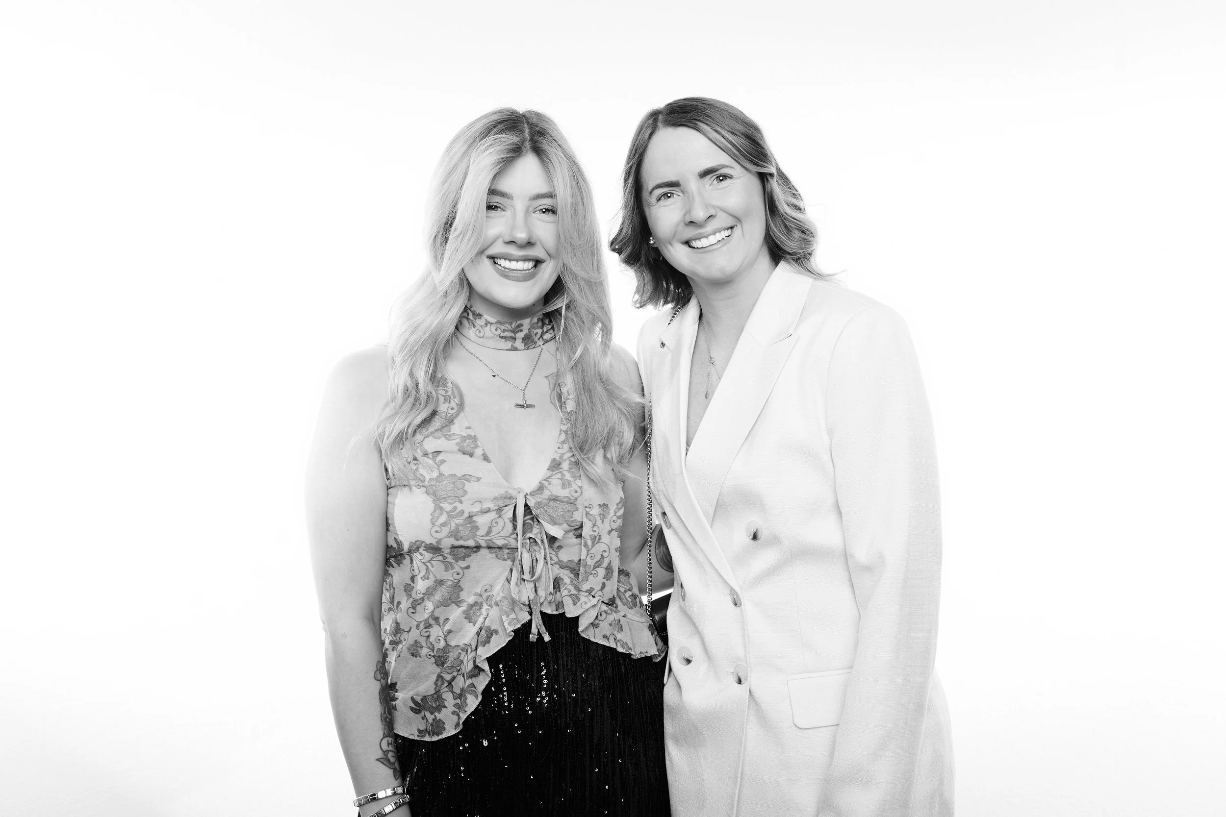 Two women standing and smiling in front of a white background, one wearing a sleeveless floral top and the other in a light-colored blazer.