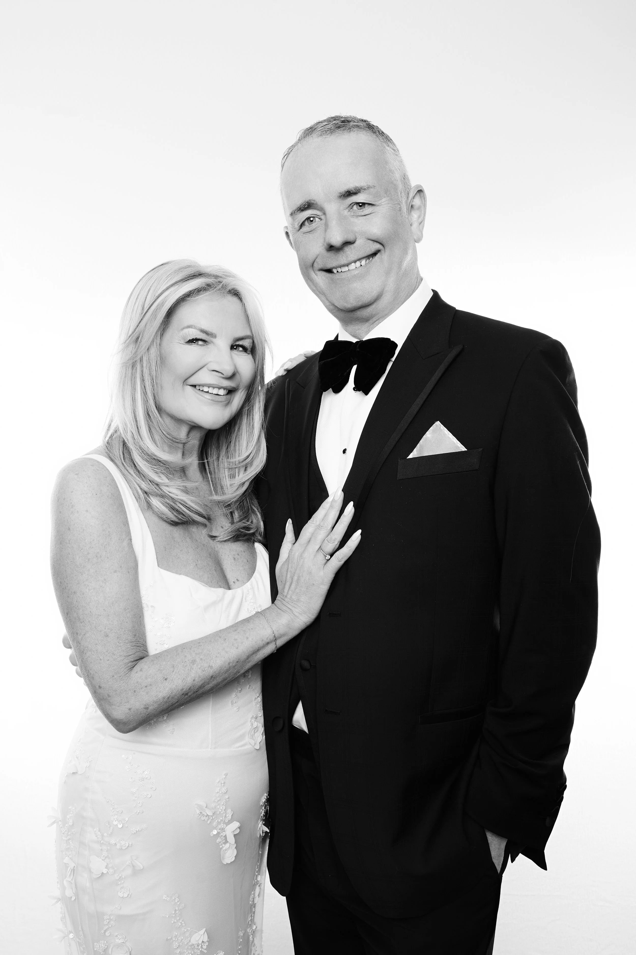 Black and white photo of a smiling woman in a white dress with floral details and a man in a tuxedo with a bow tie, both looking at the camera against a plain light background.