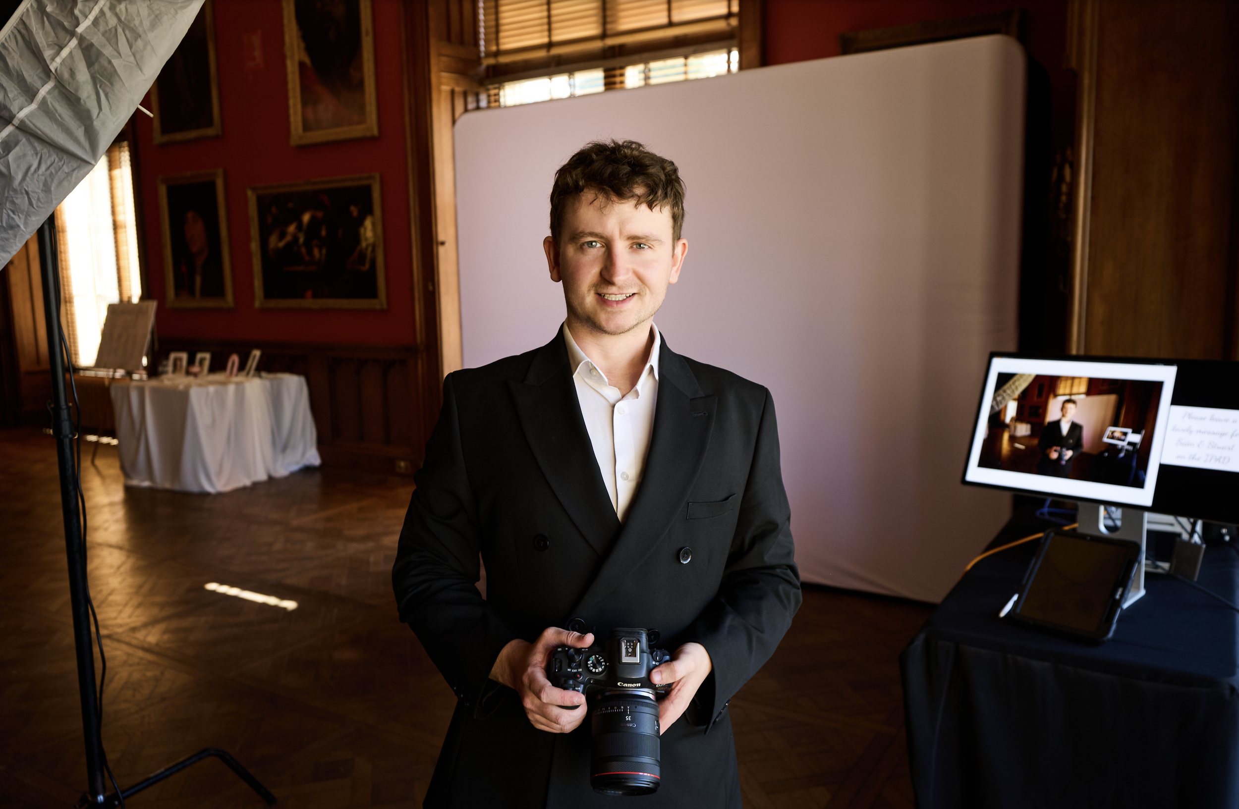 A young man in a suit holding a camera stands inside a warmly lit room with wooden paneling and framed artwork. There is photography equipment and a monitor showing a preview of the photo shoot in the background.