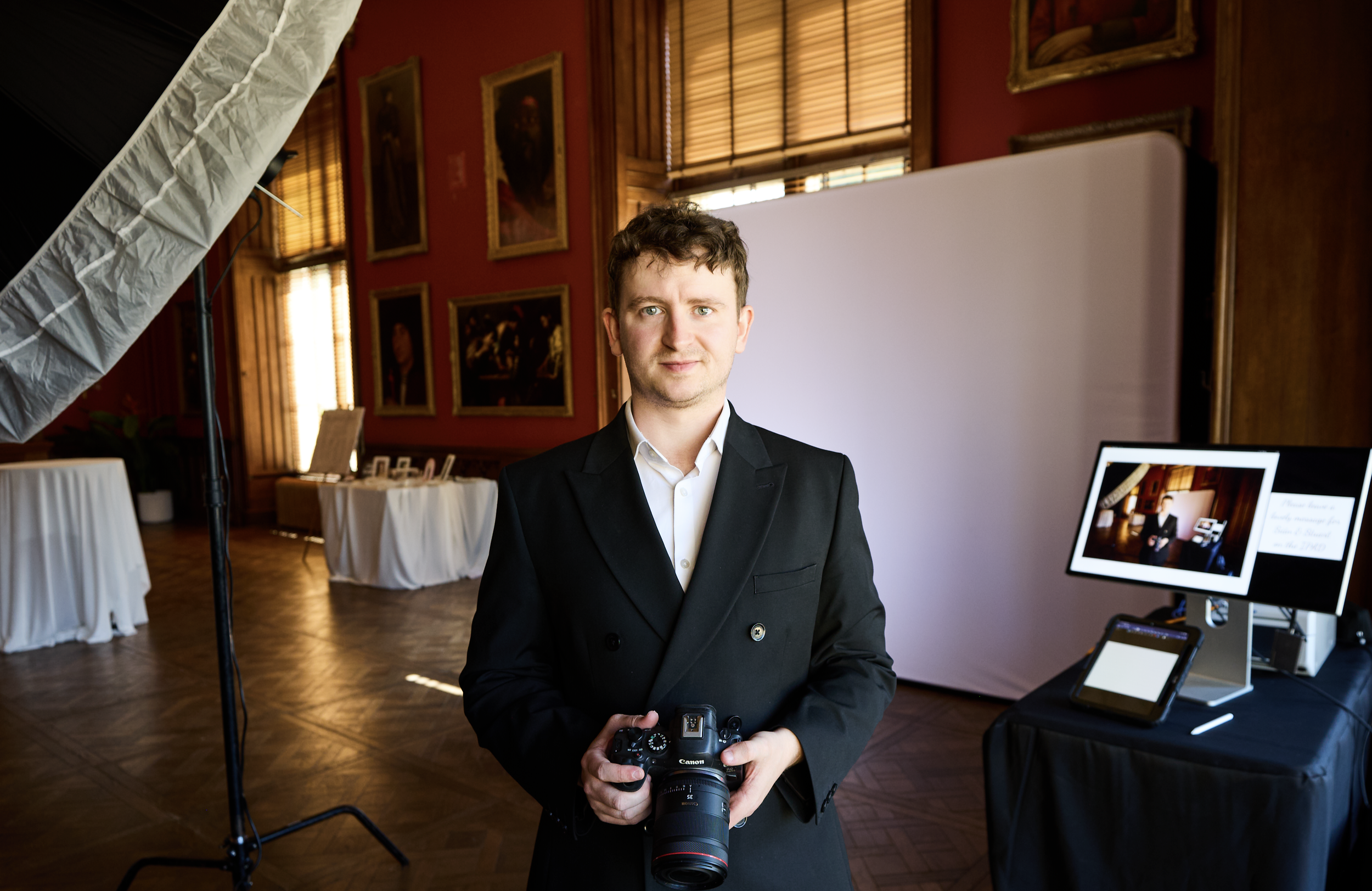 A young man in a black suit holding a Canon camera, standing in a room with red walls and framed artwork, professional photography setup with lighting and a computer monitor displaying the scene.