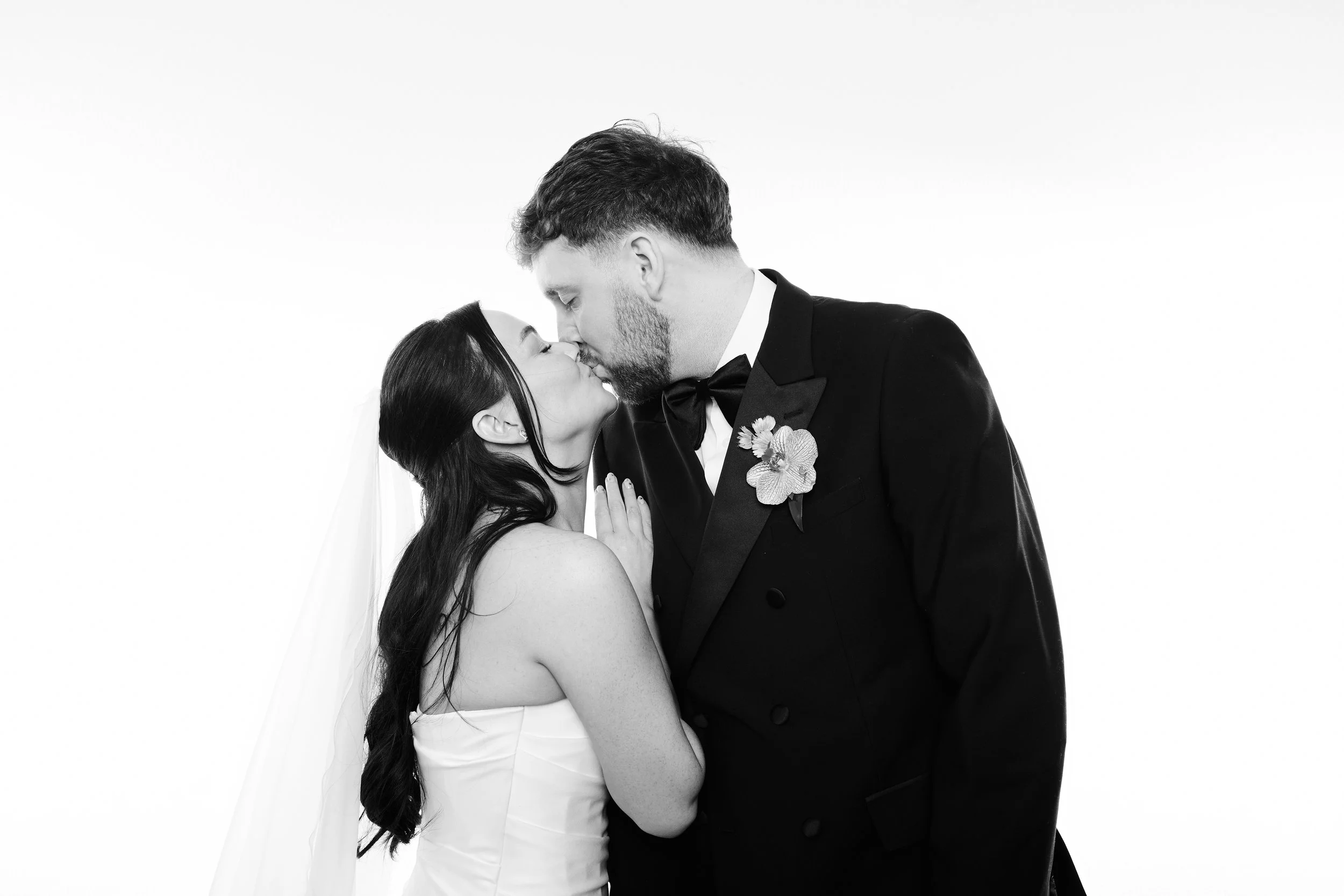 A black and white photo of a bride and groom kissing. The bride has long dark hair, and the groom has short hair and a beard. The groom is wearing a tuxedo with a boutonnière, and the bride is in a strapless wedding gown with a veil.