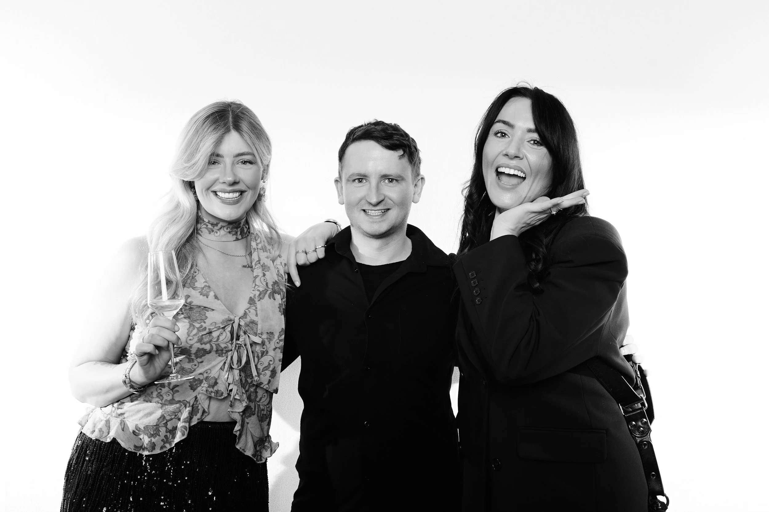 Three people, two women and one man, smiling and posing together against a plain background. The woman on the left holds a wine glass and has long blonde hair, the man in the middle has short dark hair, and the woman on the right has long dark hair a