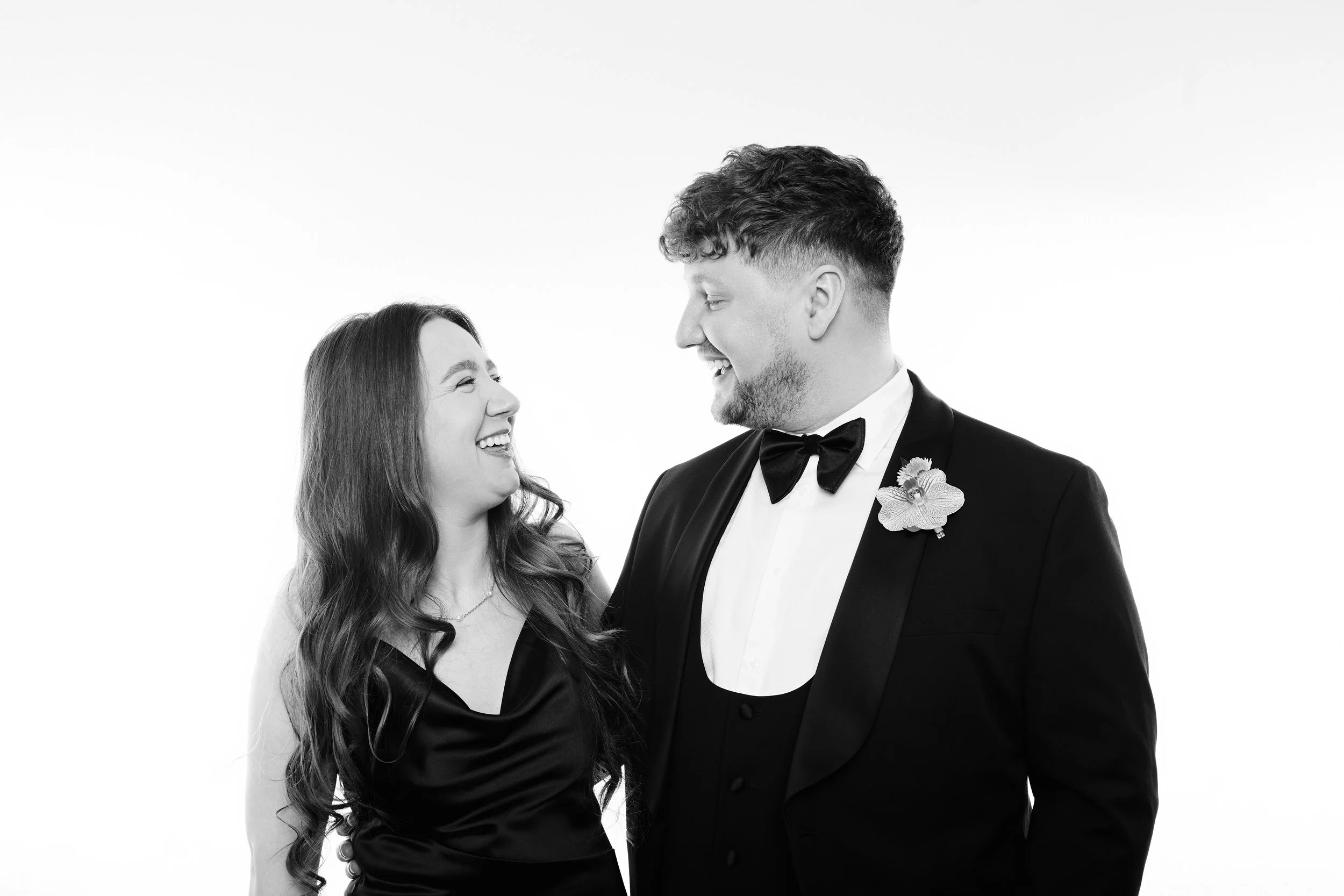A black-and-white photo of a smiling woman and man facing each other, dressed in formal attire, with a white background.