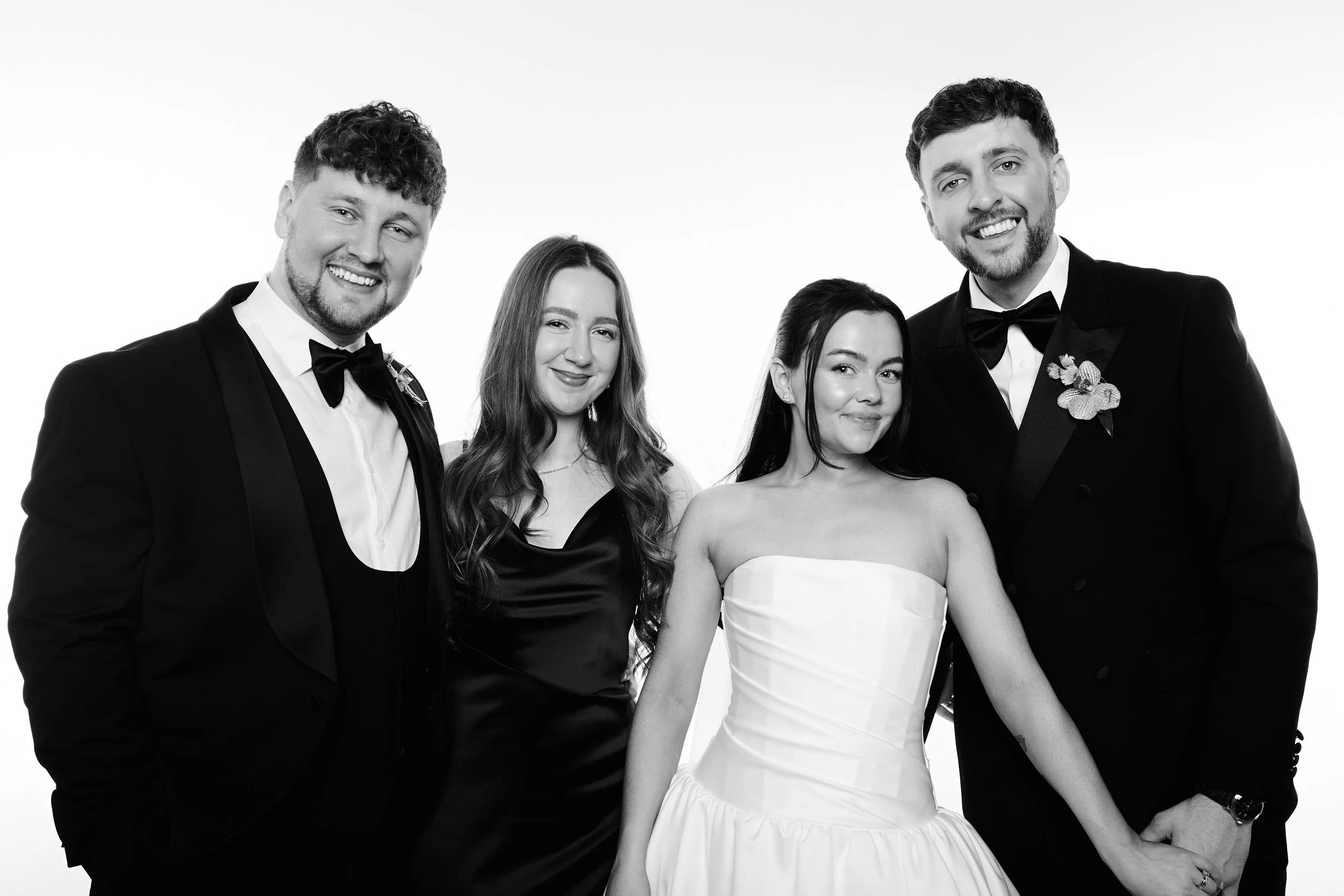 A group of five people dressed in formal attire, with two men in tuxedos and three women in elegant dresses, smiling and posing together in a studio with a white background.