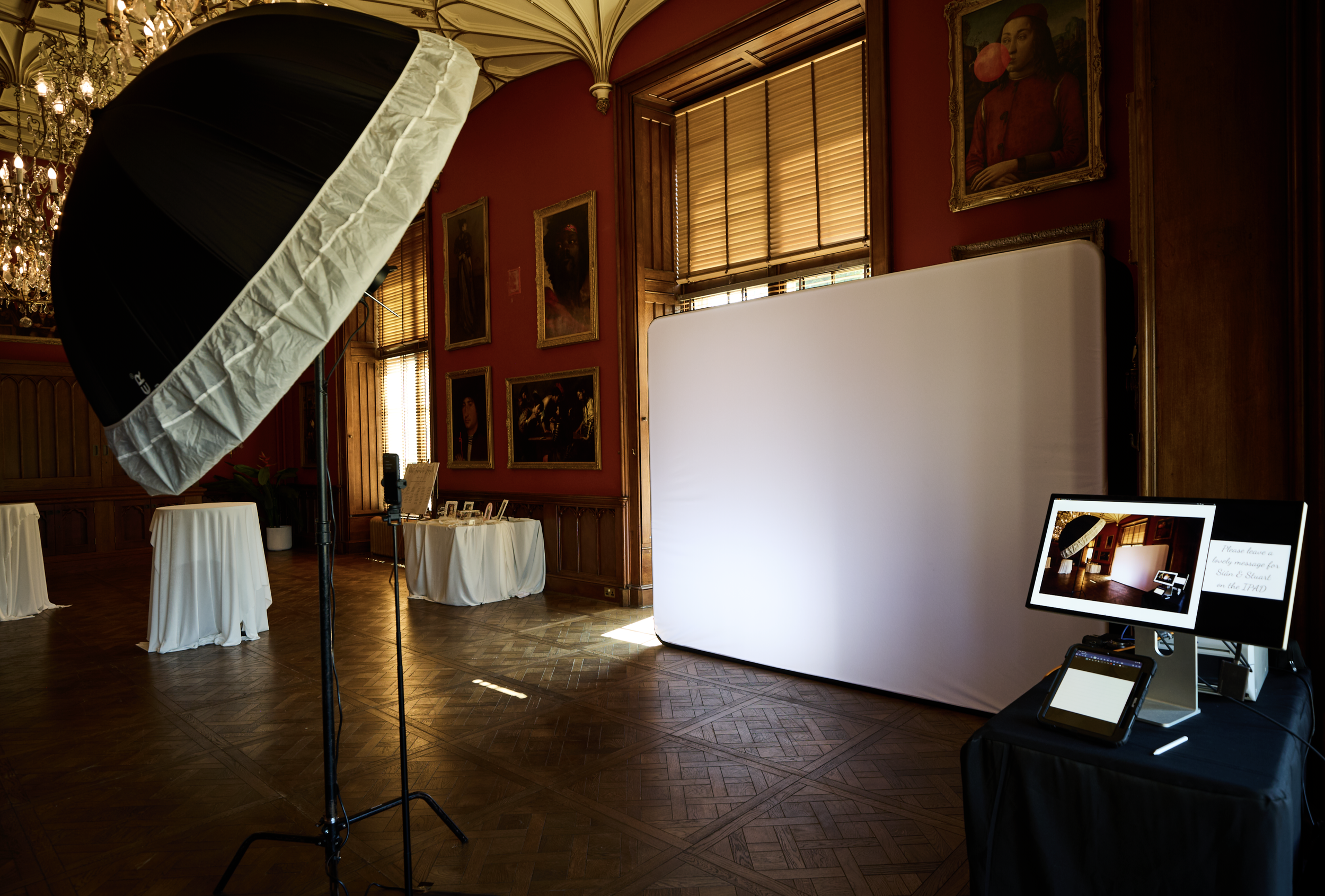 Photographer's workspace with lighting equipment, a white backdrop, and display screens in a room decorated with red walls, wooden paneling, and framed portraits. Chairs with white covers are visible, and natural light is coming through windows with wooden blinds.