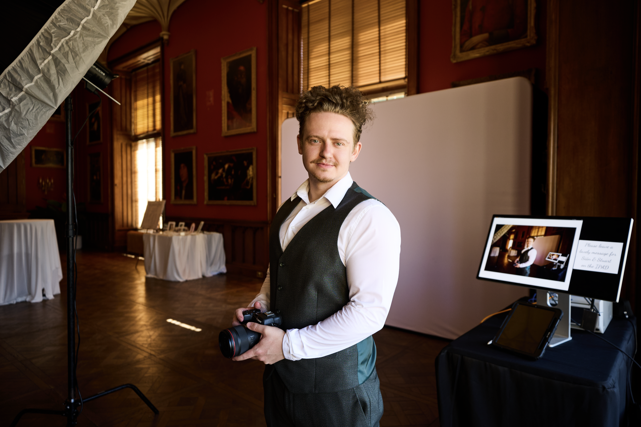 A young man in a white shirt and dark vest holding a camera stands in a room with red walls, wooden accents, and framed portraits, possibly at a photography event.