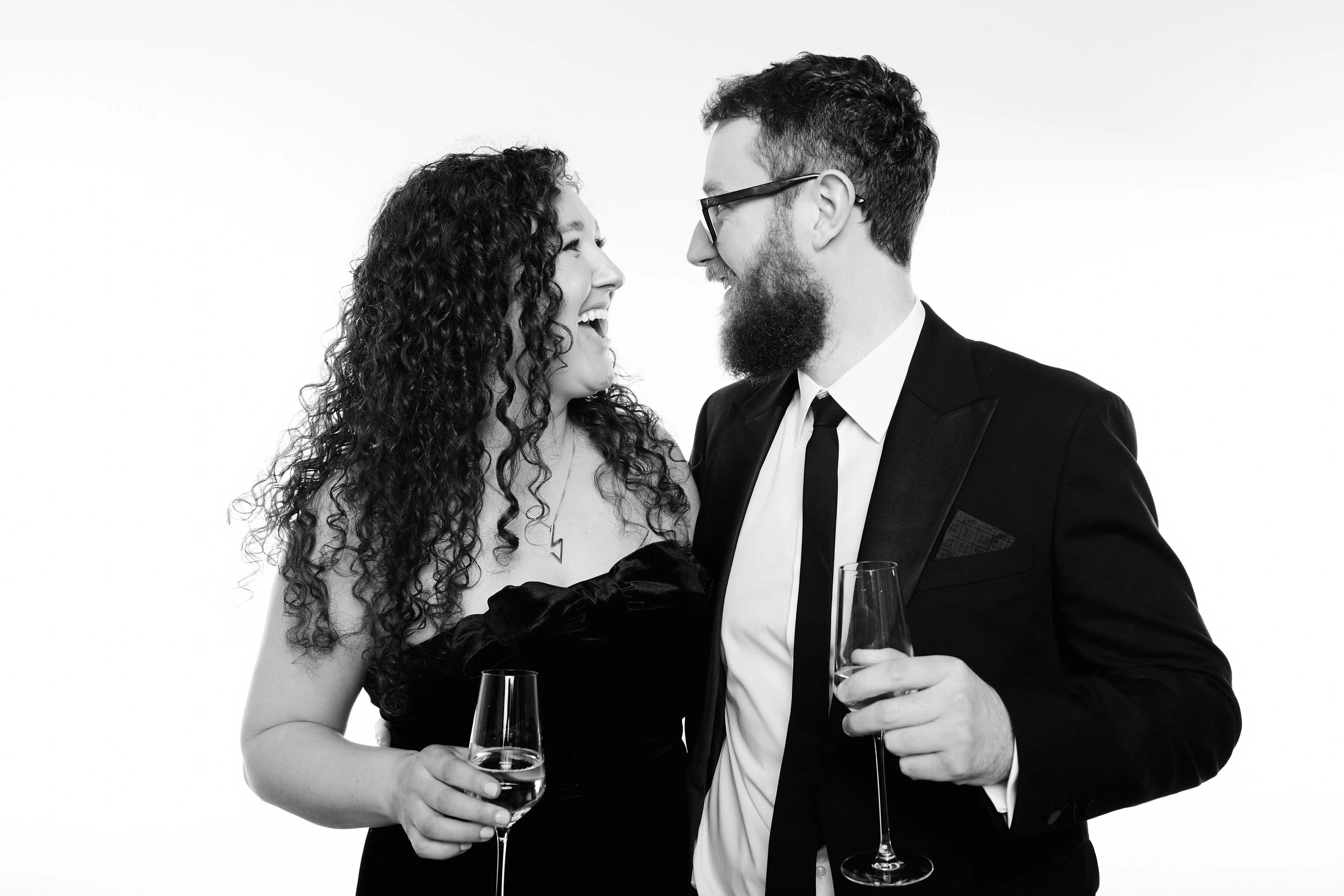 A black-and-white photo of a young woman and man at a formal event, smiling and facing each other while holding champagne glasses. The woman has curly hair and is dressed in a strapless dress with gloves, and the man has glasses, a beard, and is wear