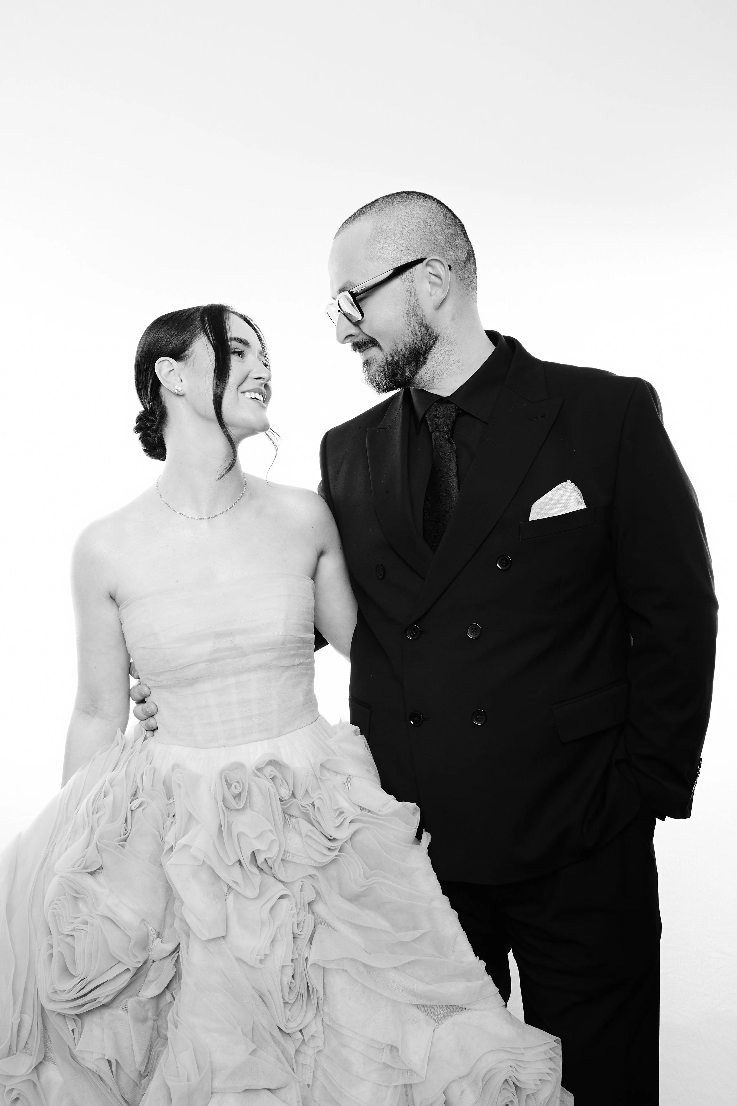 A black-and-white photo of a woman in a wedding dress and a man in a suit looking at each other and smiling.