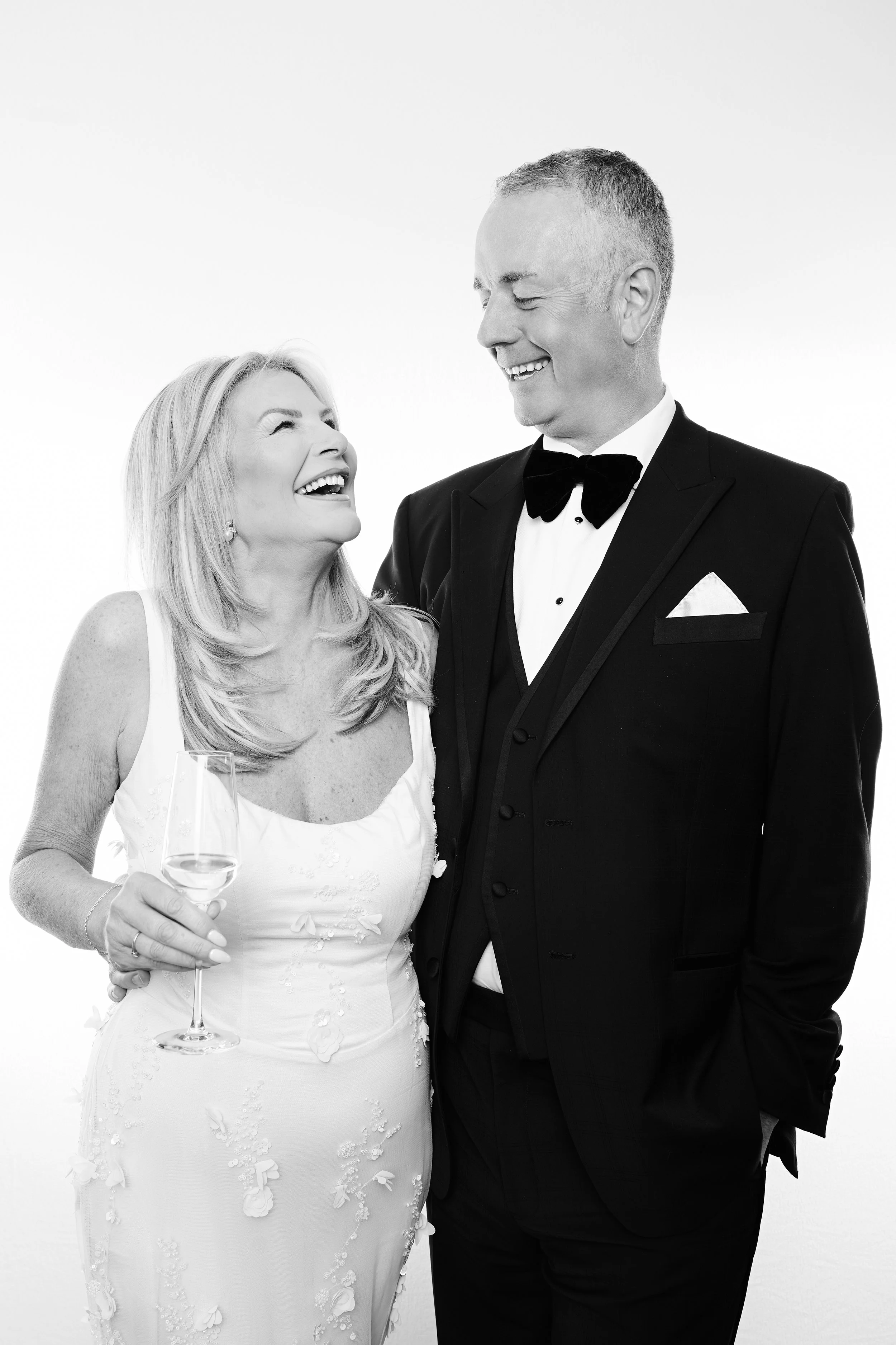 A smiling woman in a wedding dress holding a glass of wine, looking up at a smiling man in a tuxedo, both appearing joyful and happy.