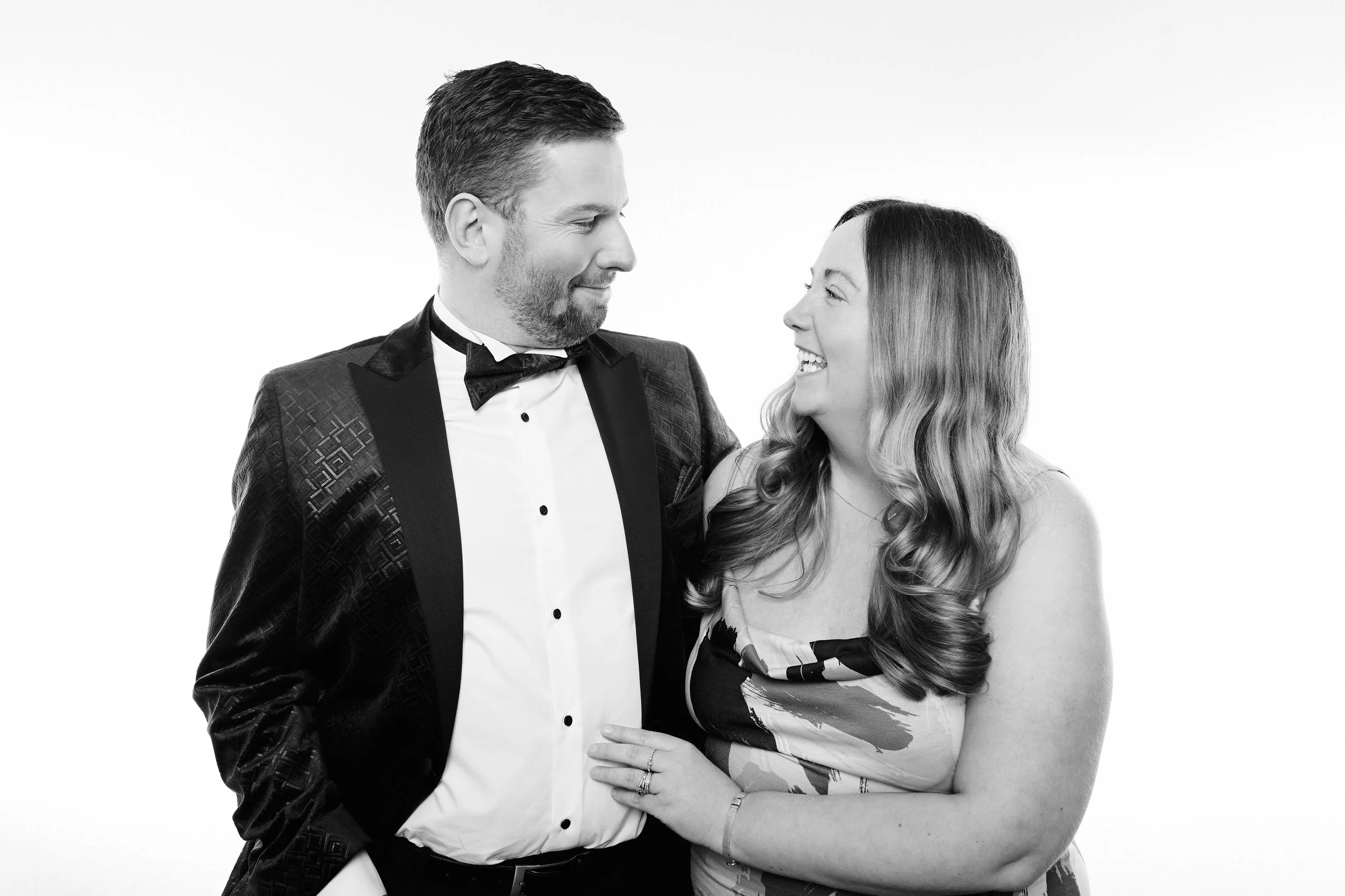 A black-and-white photo of a man and woman smiling at each other, dressed in formal attire, with the woman wearing a patterned dress and the man in a tuxedo.