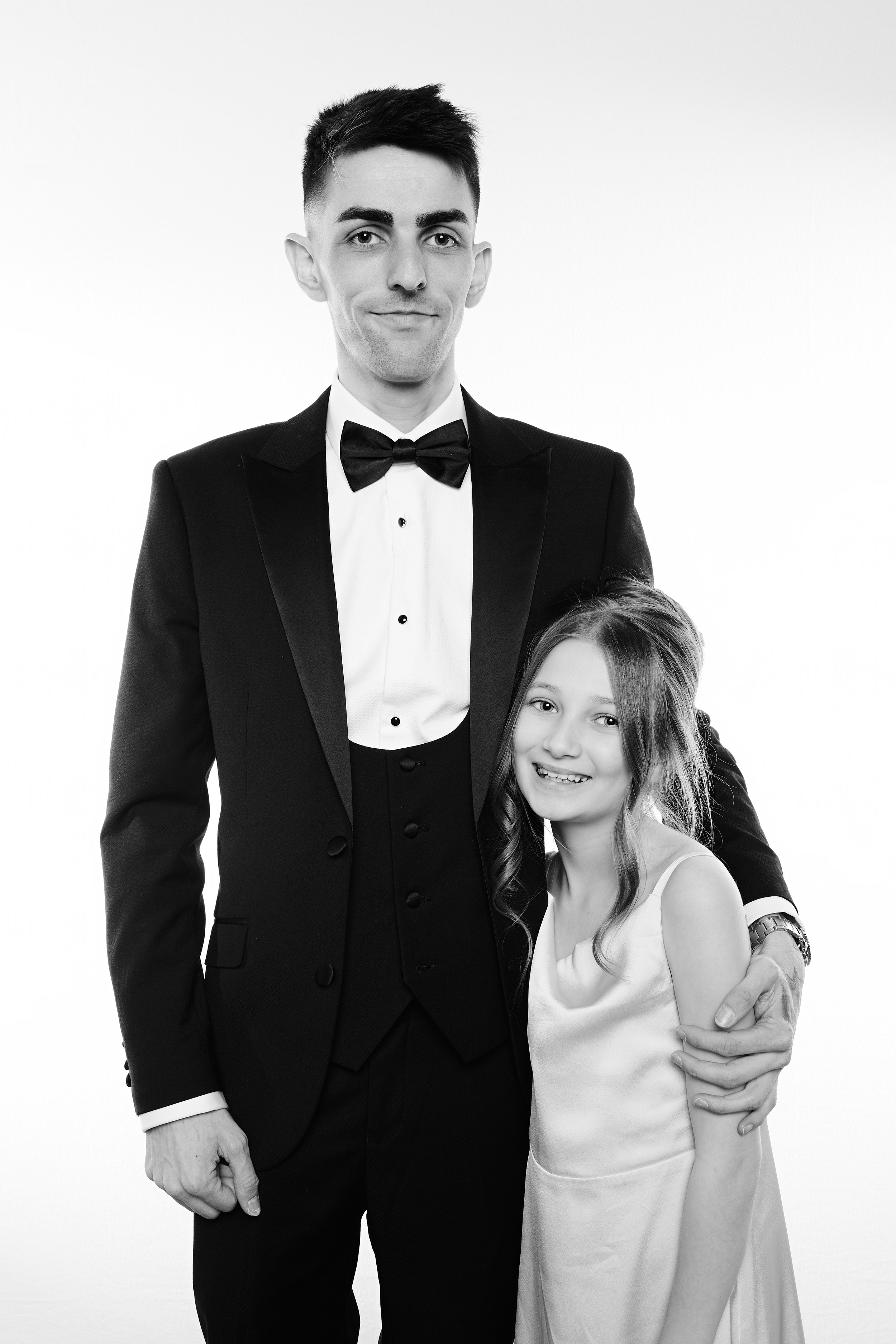 A man in a tuxedo and a young girl in a dress standing together, smiling, in a black-and-white photo.