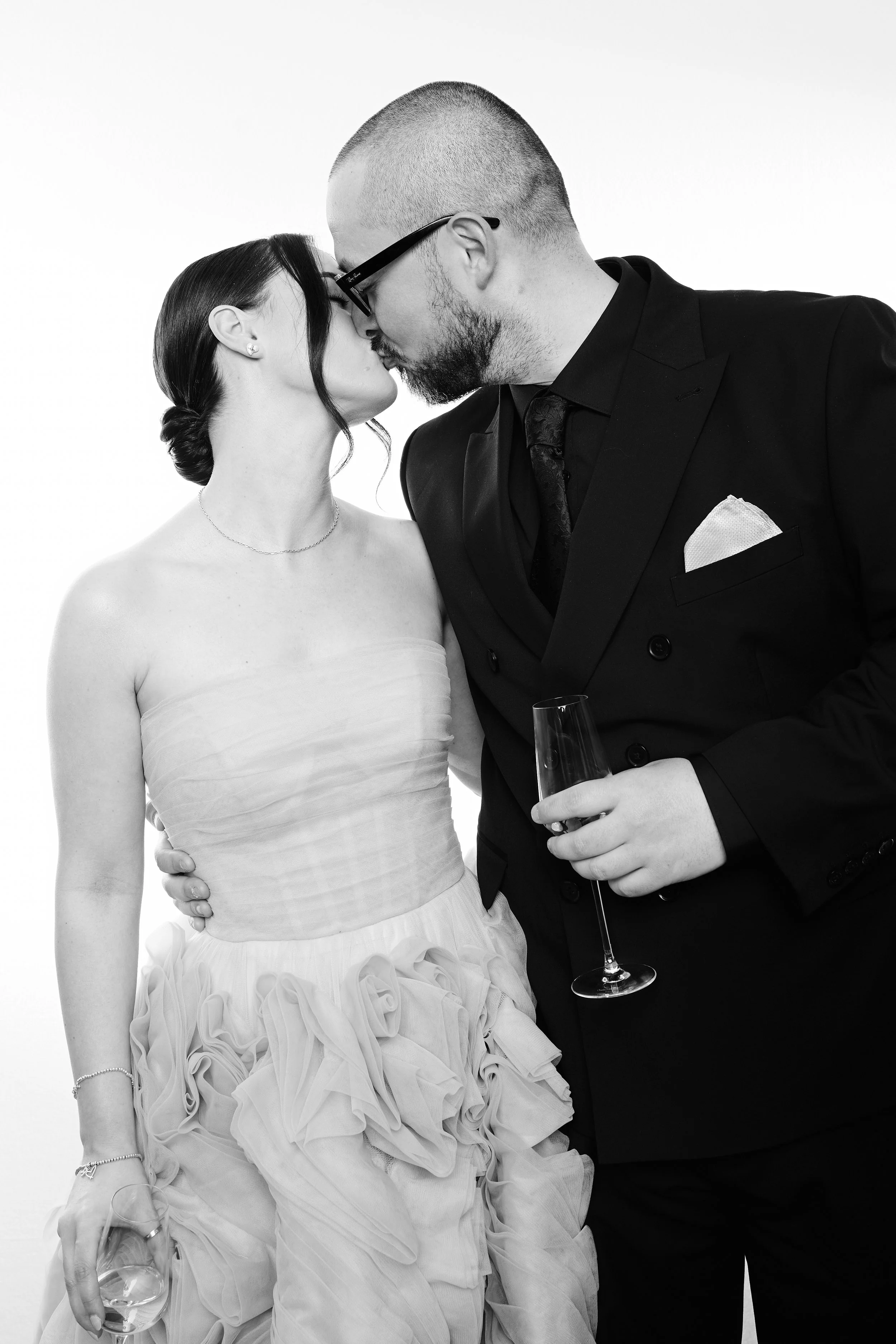A black and white photograph of a couple sharing a kiss, with the woman in a strapless dress and the man in a dark suit, holding a glass of champagne.