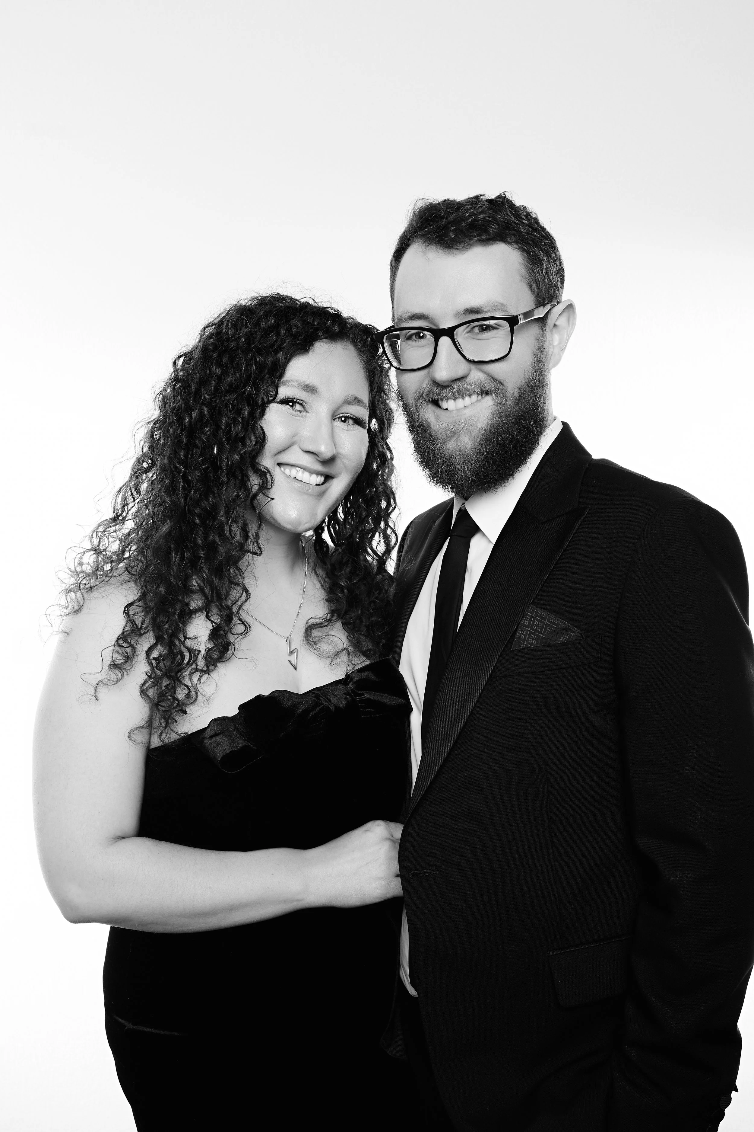 A black and white photo of a smiling woman with curly hair and a man with glasses and a beard, both dressed in formal attire, standing close together.