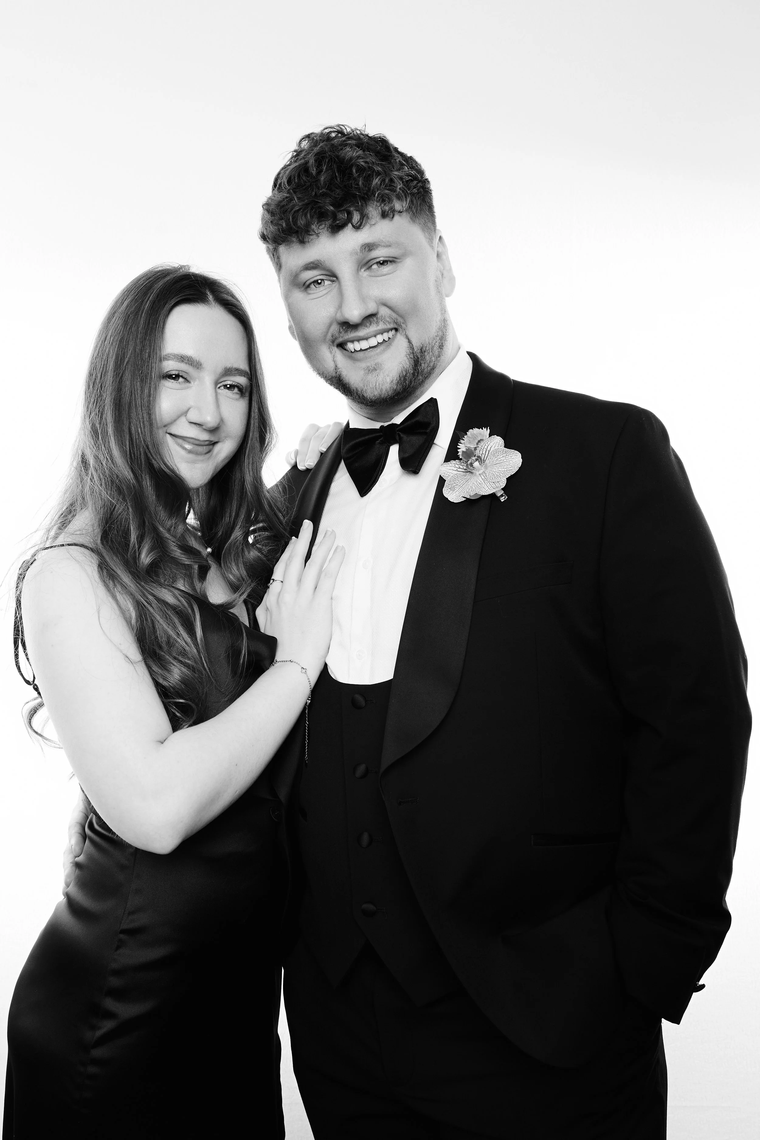 Black and white photo of a smiling couple dressed in formal wedding attire. The woman has long wavy hair and is gently touching the man's chest. The man is wearing a tuxedo with a bow tie and a flower boutonniere.