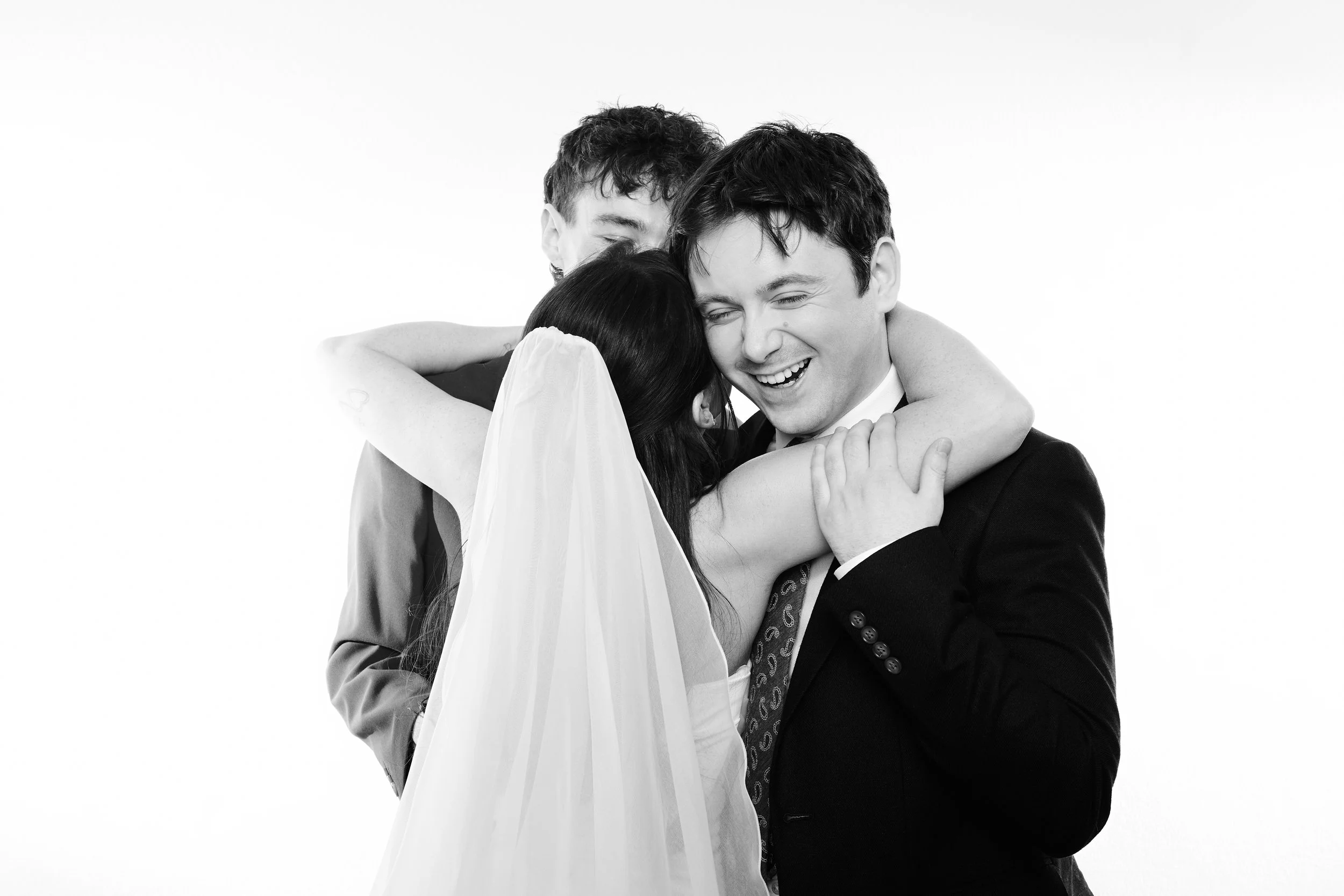 Three people hugging, one appears to be a bride in a dress and veil, the others are men in suits, all smiling.