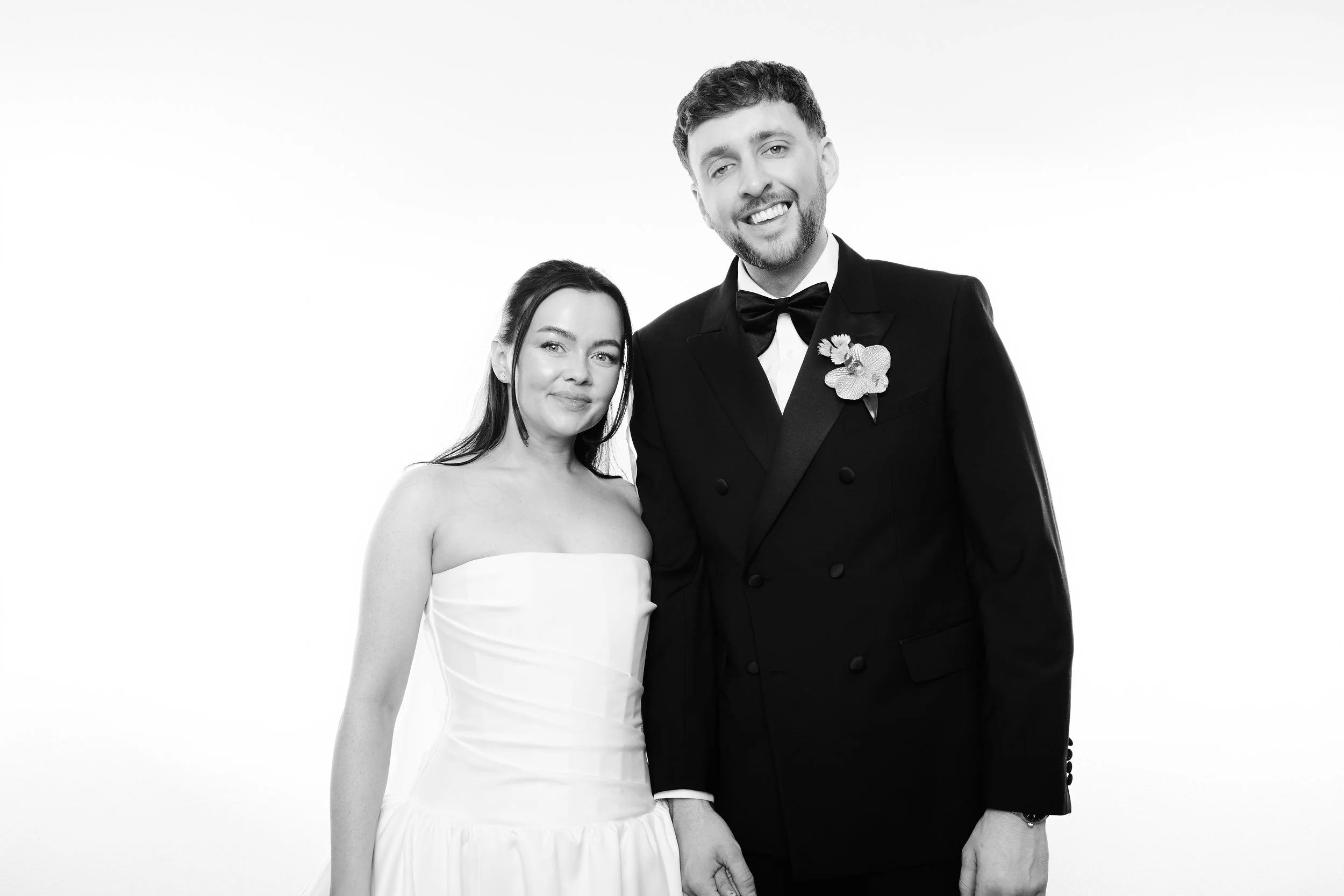 A black-and-white photo of a smiling woman in a strapless dress standing next to a man in a tuxedo with a boutonniere, set against a plain background.