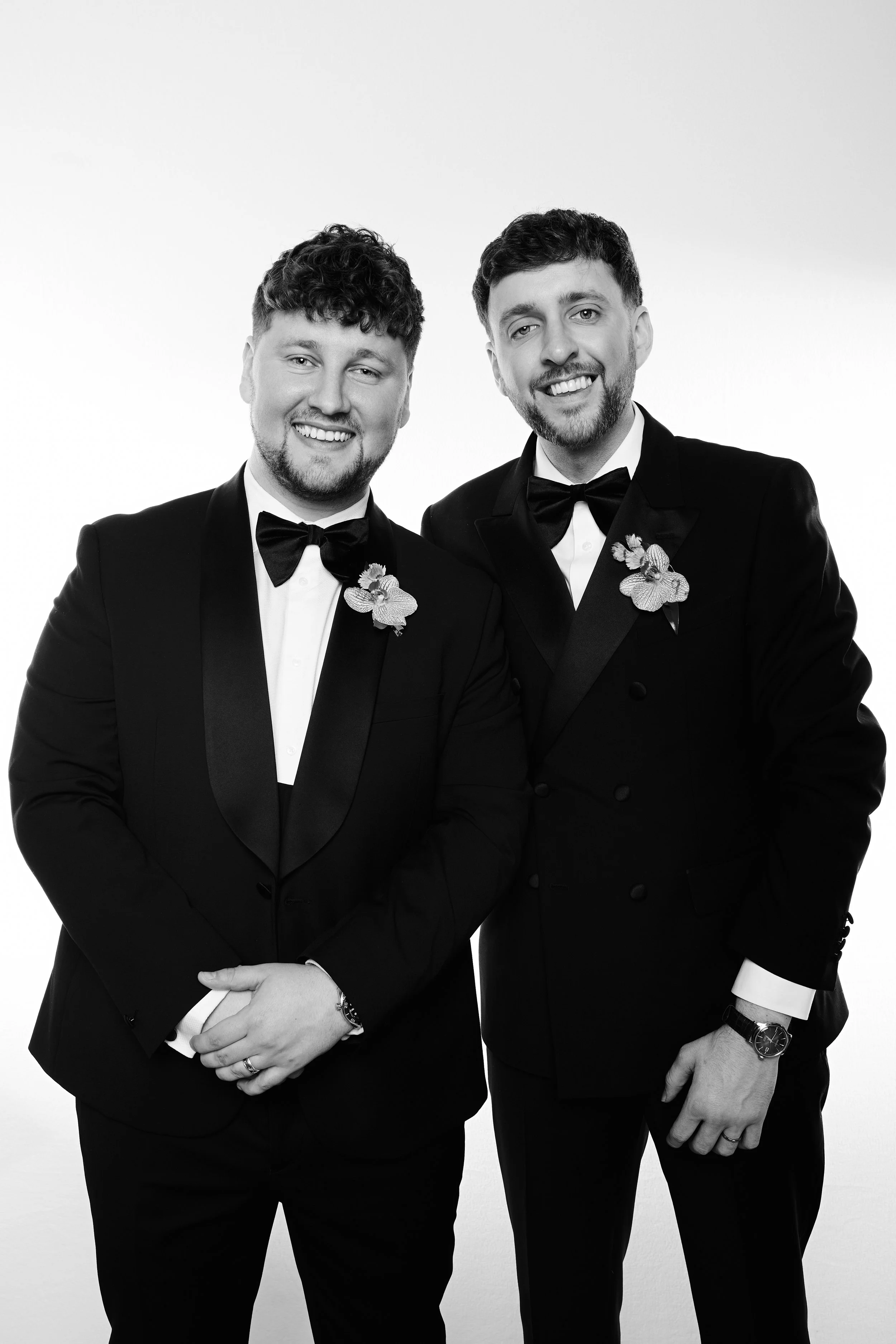 Two men dressed in black tuxedos with bow ties and orchids on their lapels, smiling and standing close together against a plain background.